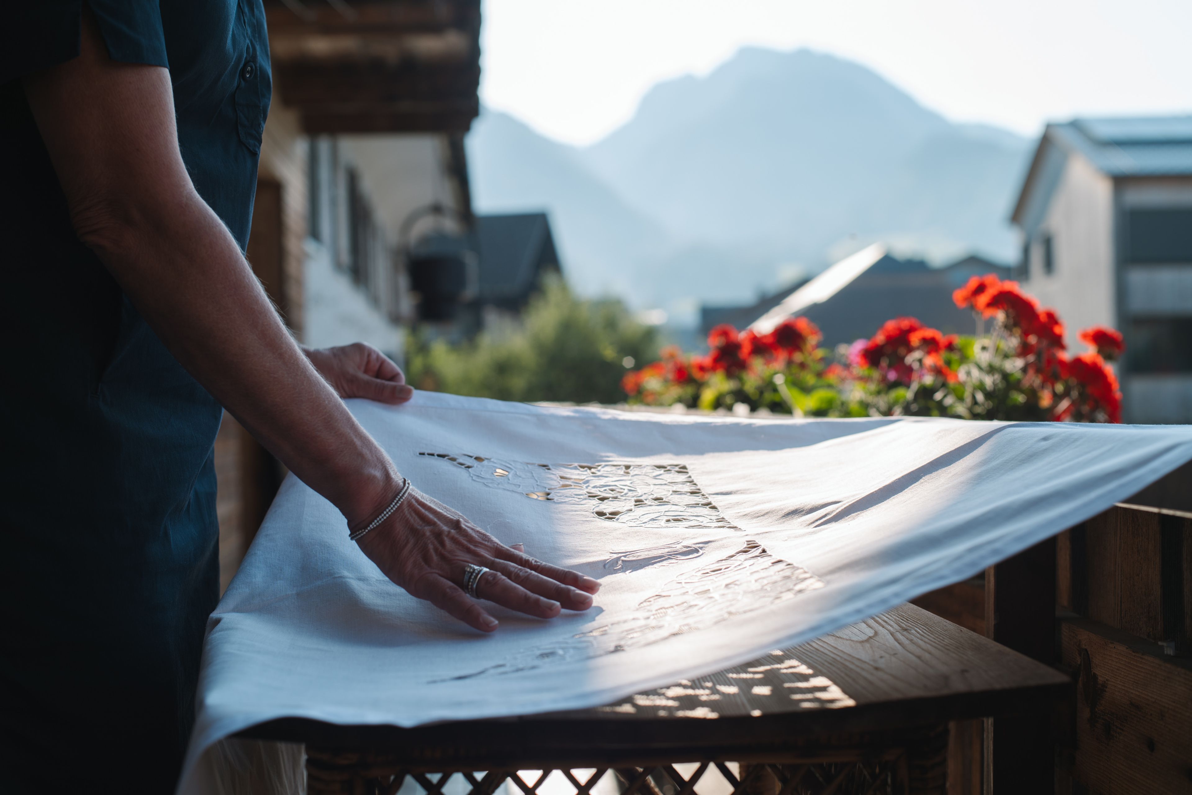 Person drapes a linen cloth over a wooden railing, with a mountain view and red flowers in the background.