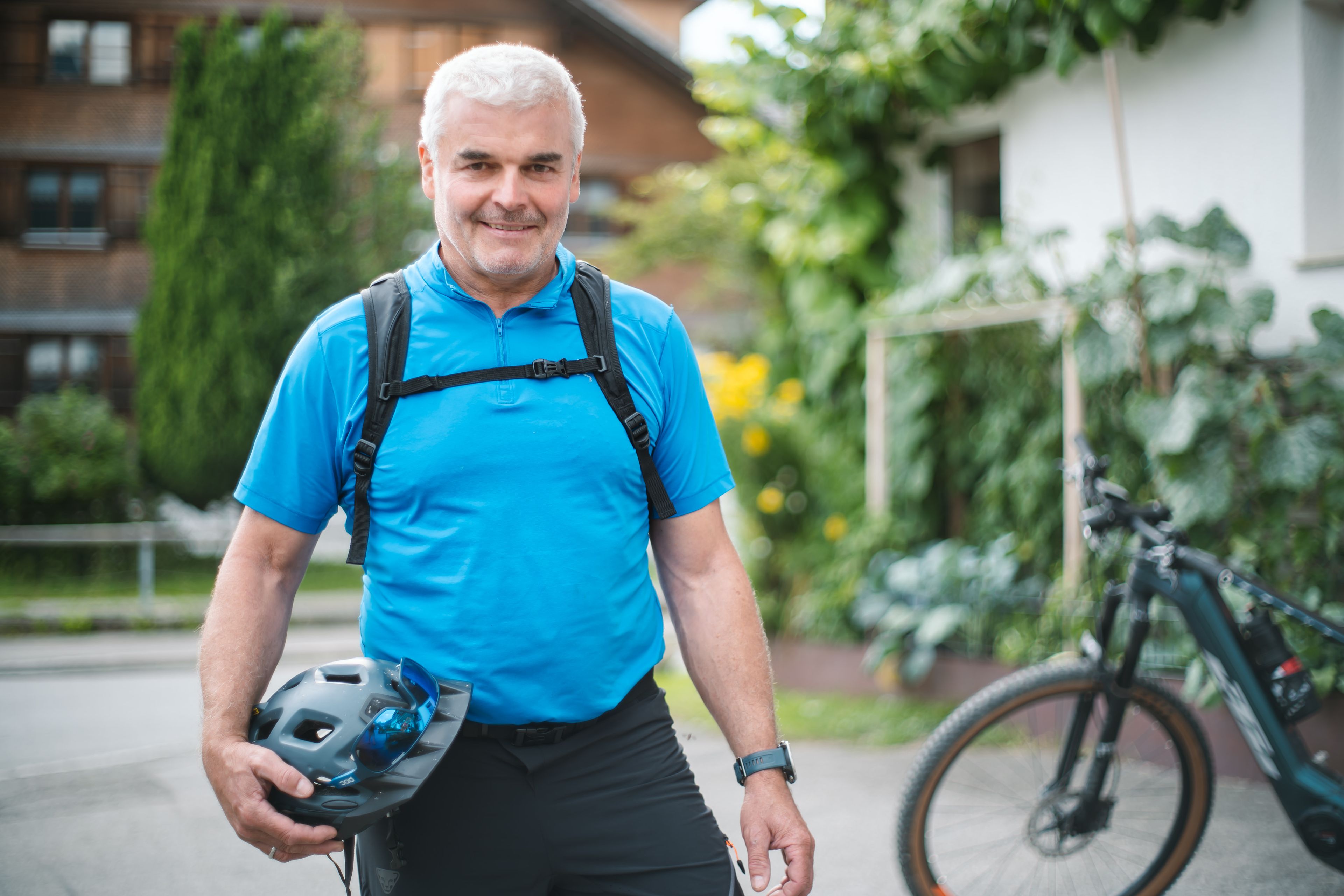Mann in blauem Shirt hält Fahrradhelm, steht mit Fahrrad im Grünen vor einem Haus, lächelt freundlich.