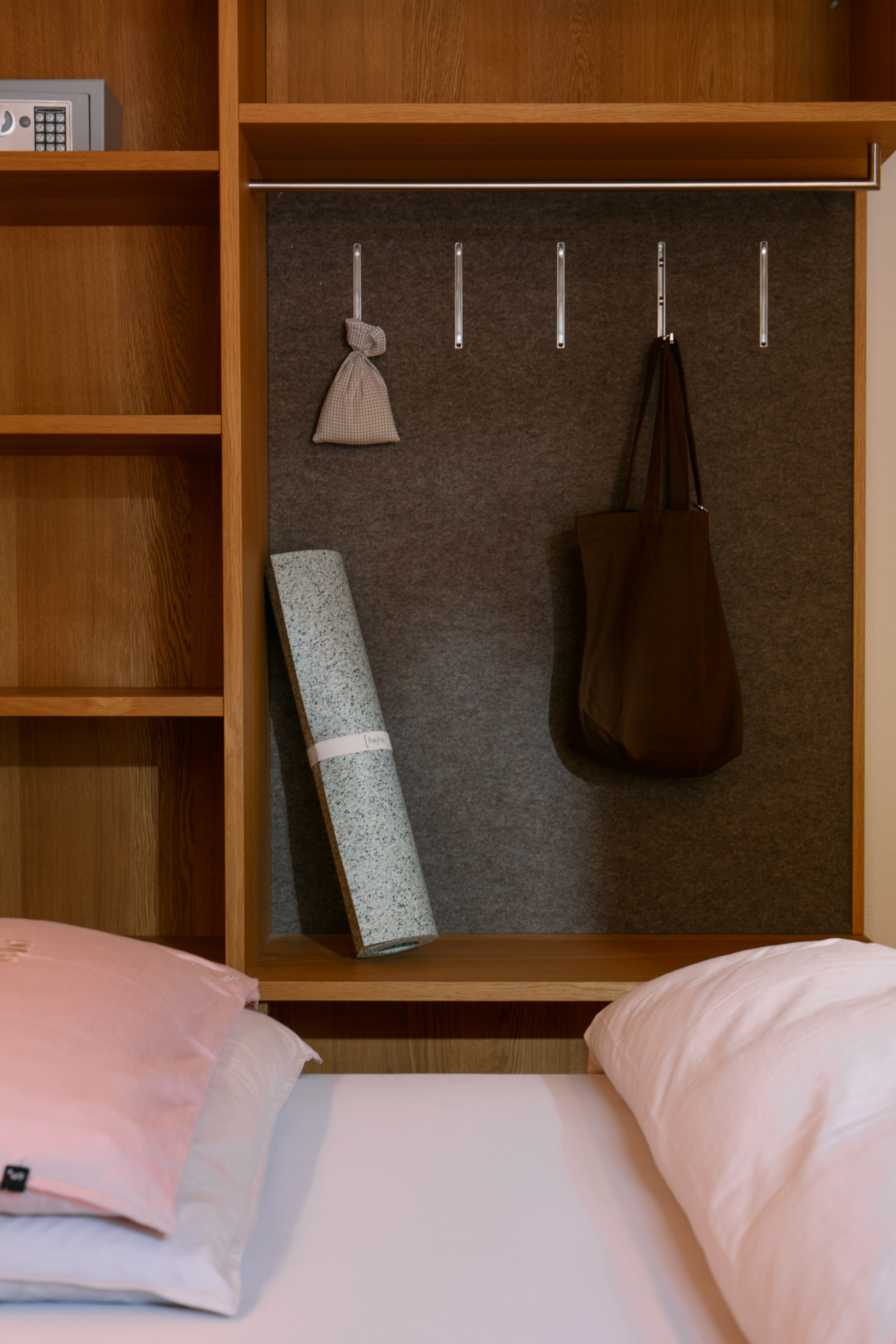 Schranknische mit grauem Filzhintergrund, Haken, brauner Tasche, gerollter Yogamatte, darüber Regal mit Safe, davor Bett mit rosa Kissen.