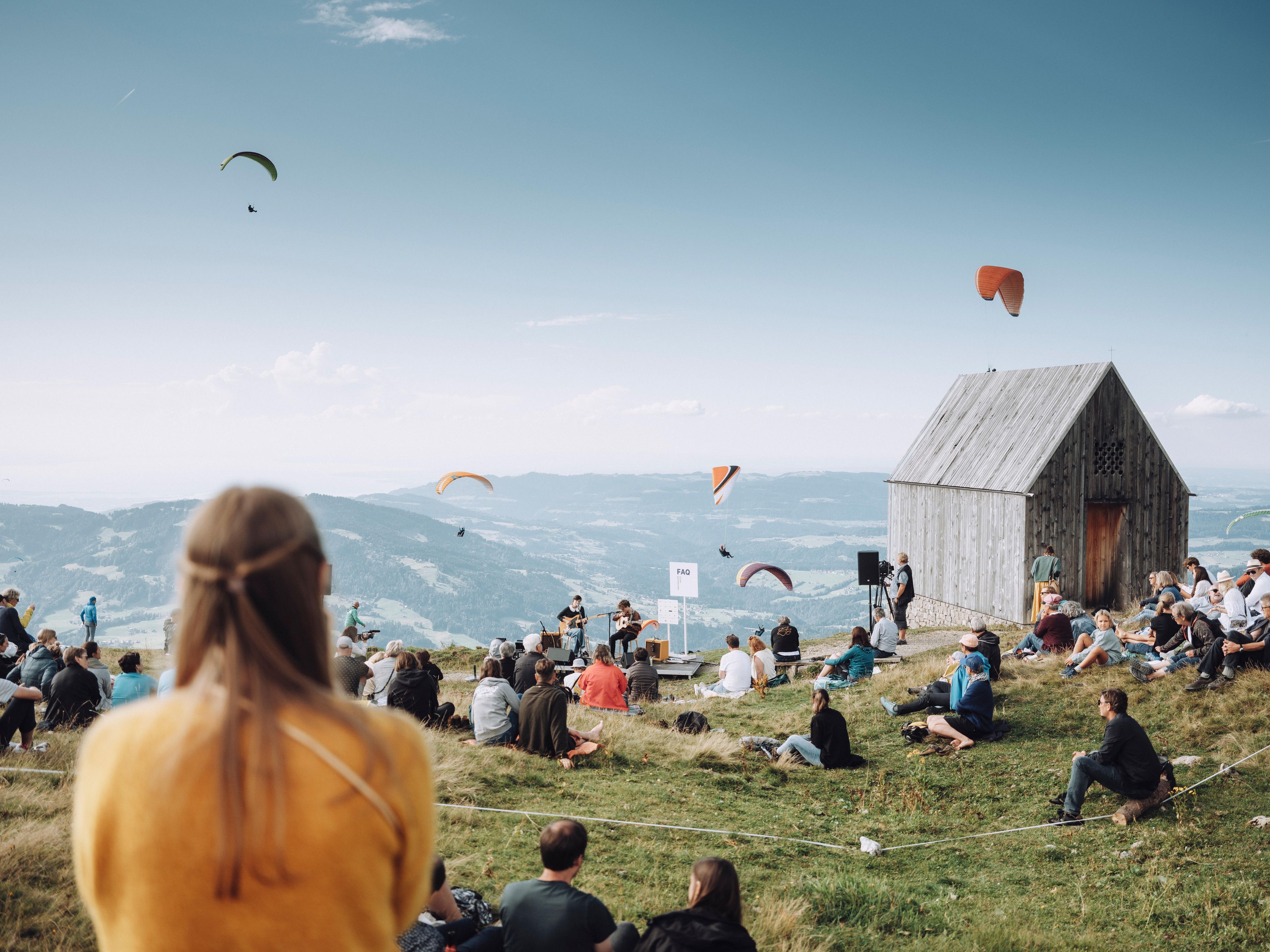 Menschen auf einem Berg mit Kapelle, die einem Konzert zuhören und Paragleiter im Hintergrund