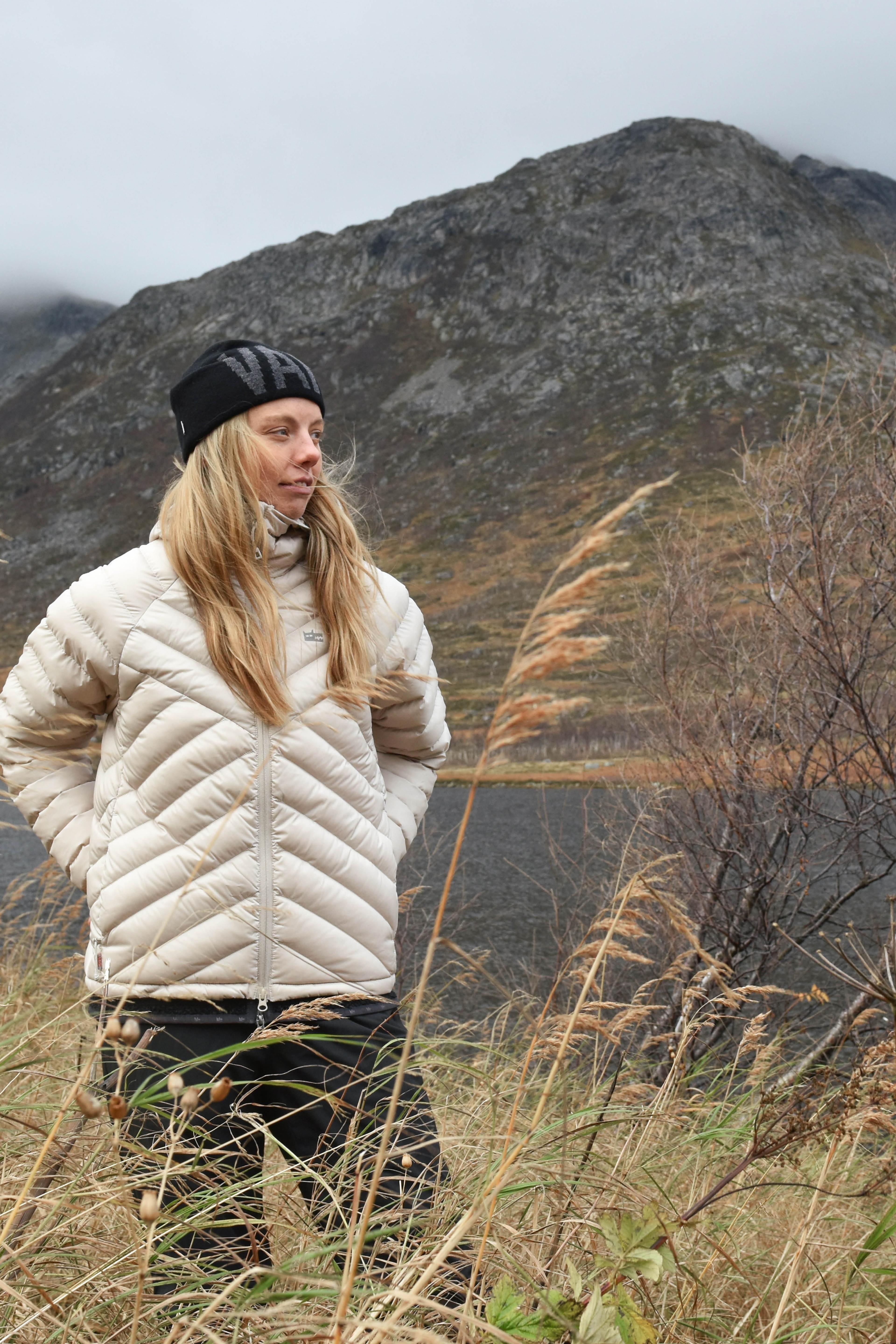 A person in a white down puffer jacket and black beanie stands in tall grass with a mountainous landscape in the background.