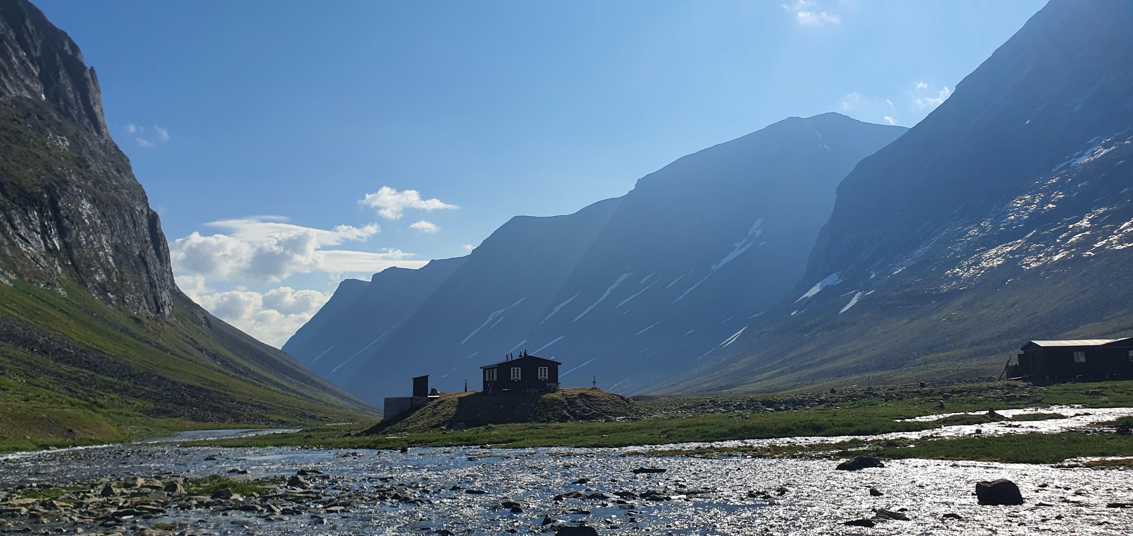Mountain valley Kebenekajse with a small cabin beside a flowing river, surrounded by steep, sunlit cliffs under a clear blue sky.