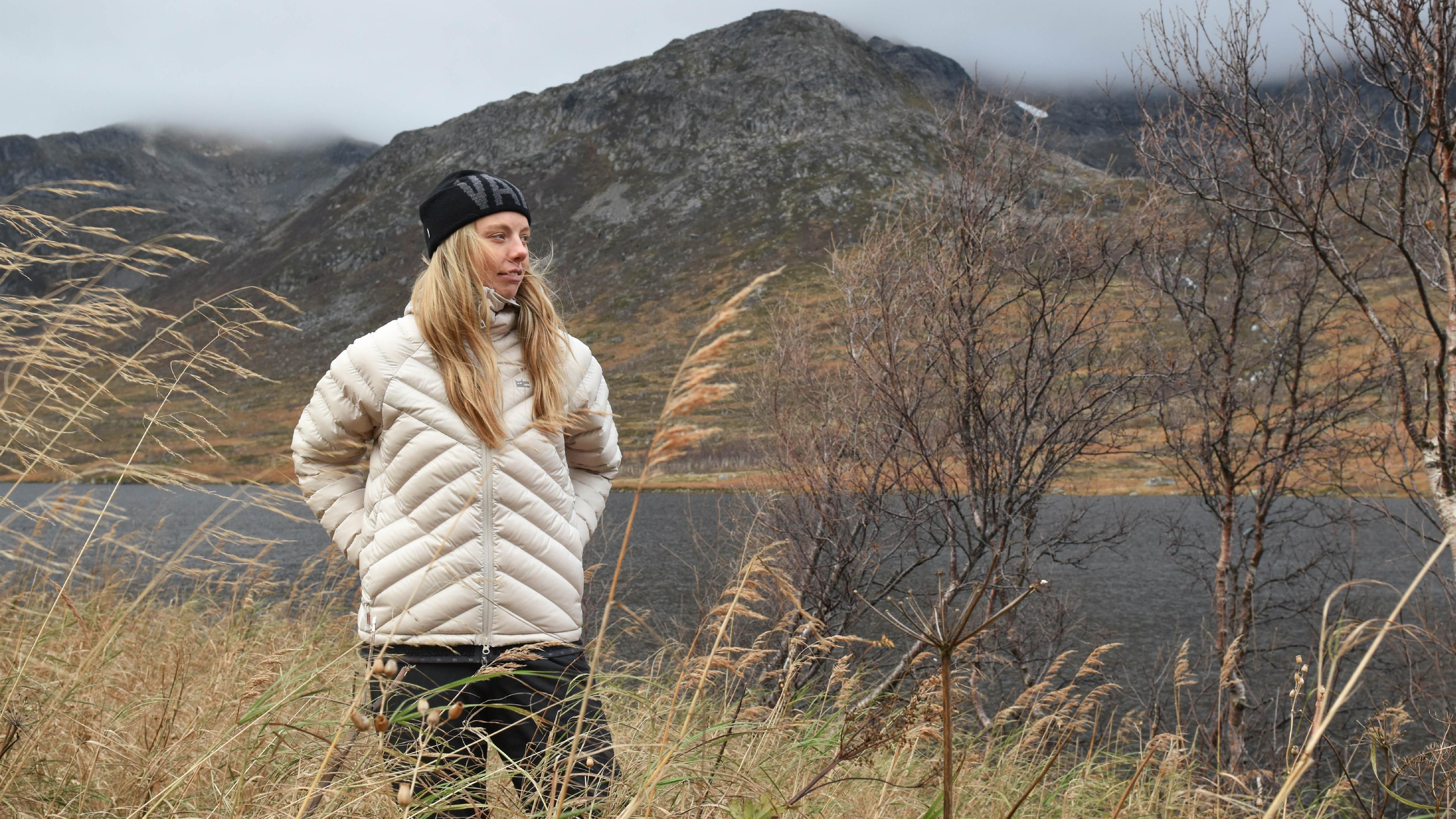 A woman in a quilted down jacket and beanie stands in tall grass beside a lake, with a mountain shrouded in clouds in the background.