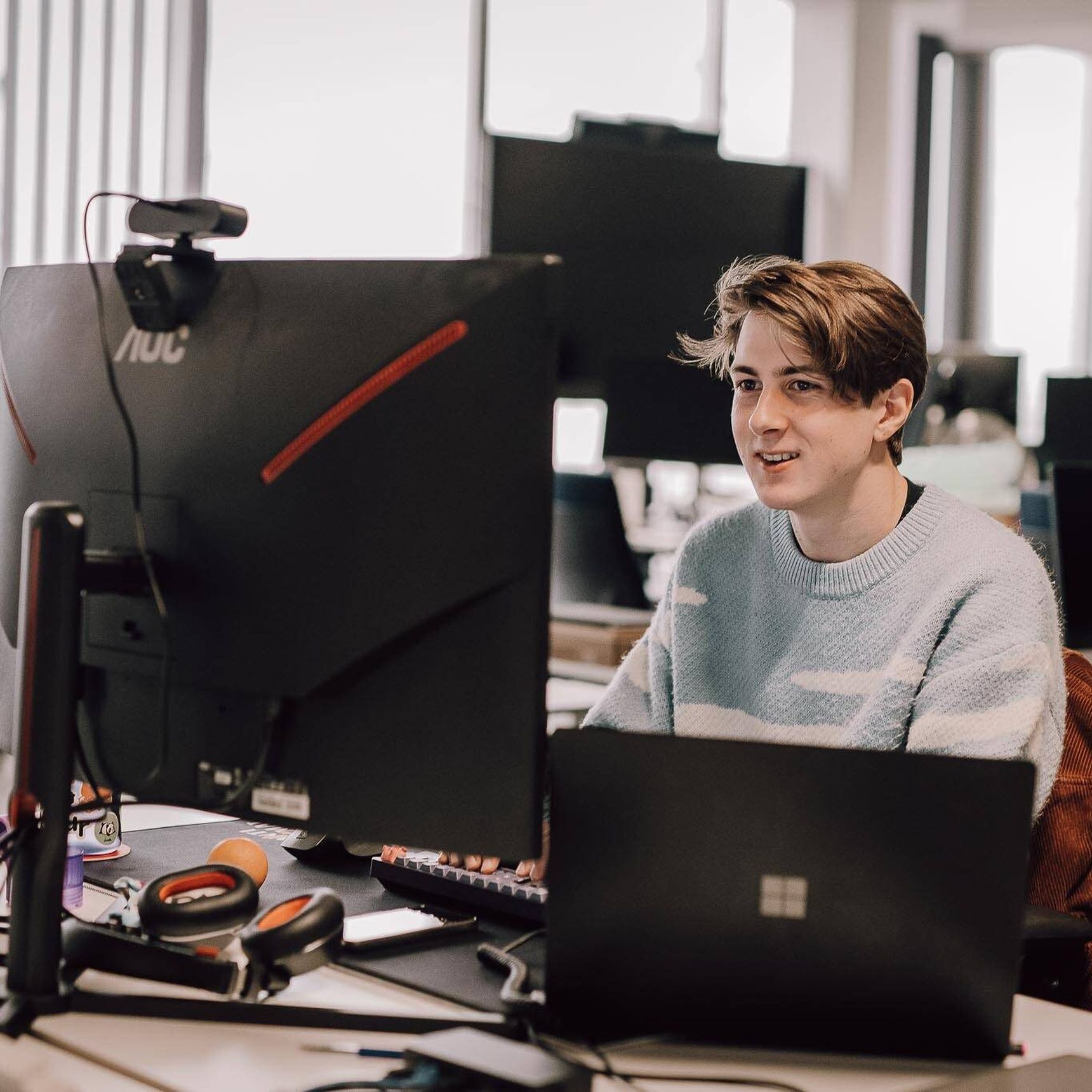 Jonge man werkt achter een computer met meerdere schermen in een modern kantoor. Hij draagt een lichtblauwe trui en lijkt gefocust.