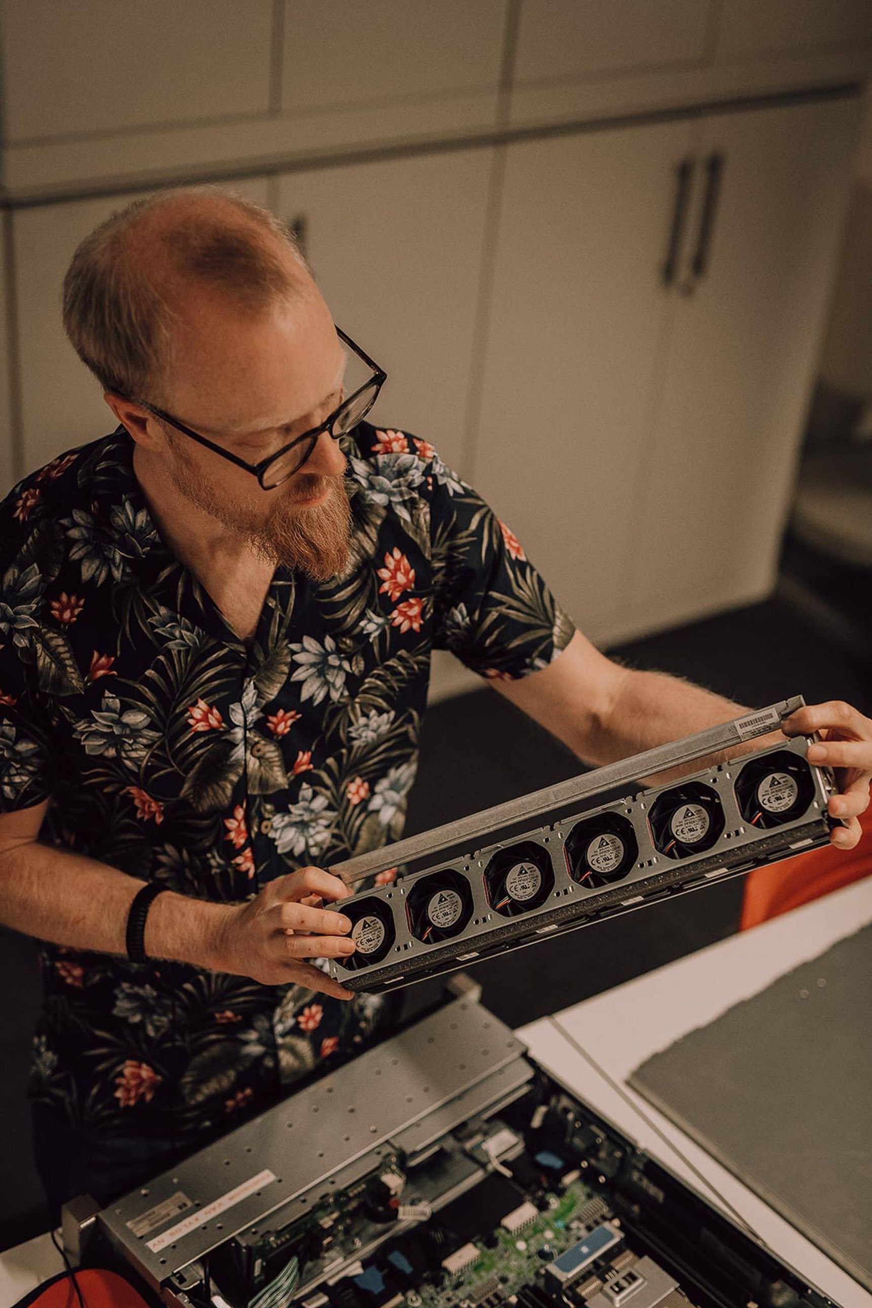 Man met bril en bloemenhemd bekijkt een computeronderdeel ventilatoren, staand boven een open computerbehuizing.