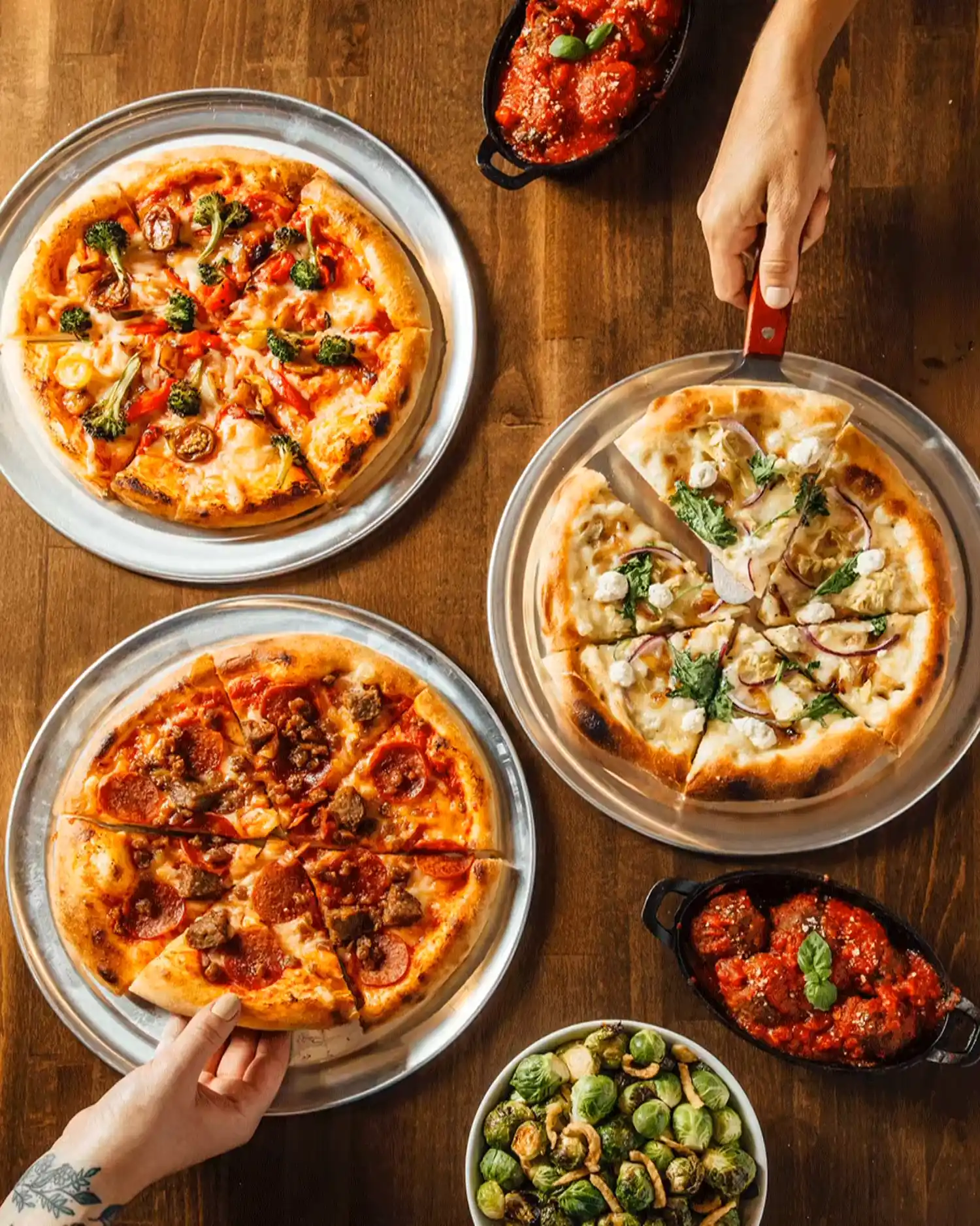 Three pizzas with various toppings on metal trays, two dishes of marinara sauce with meatballs, and a bowl of Brussels sprouts on a wooden table.