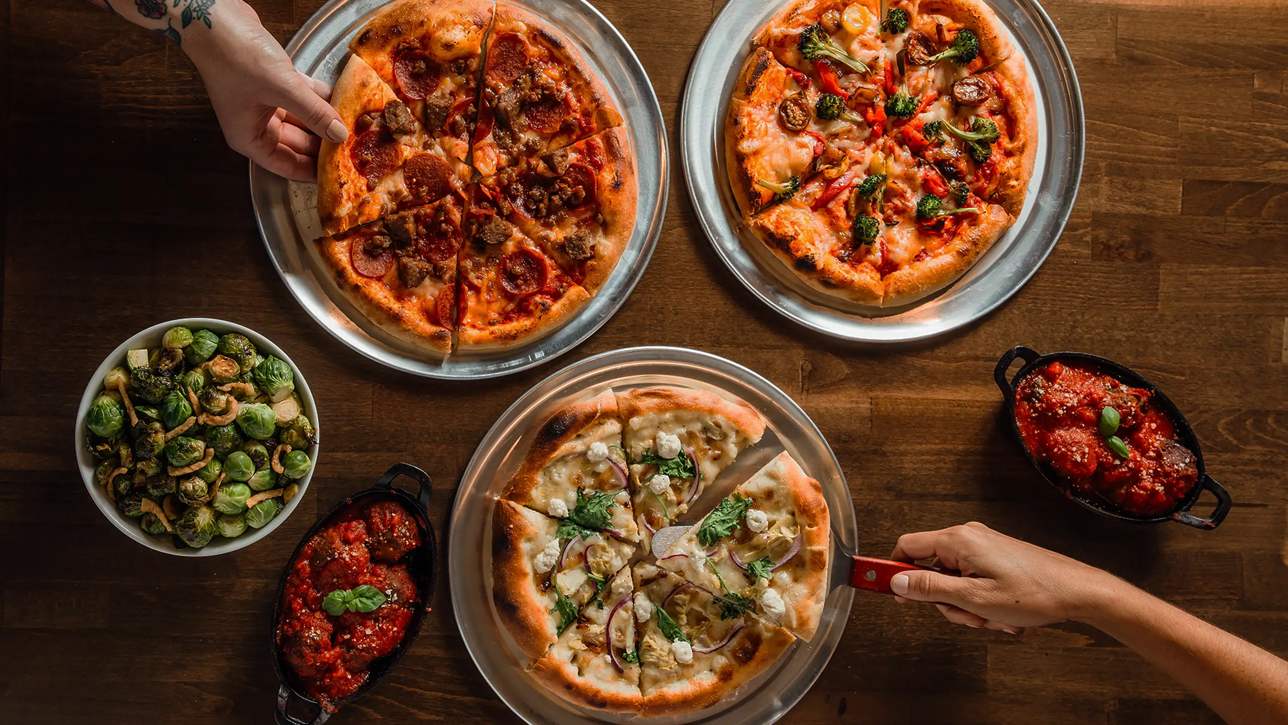 Variety of Wild Pie pizzas on metal trays with side dishes of Brussels sprouts and meatballs in tomato sauce on a wooden table.
