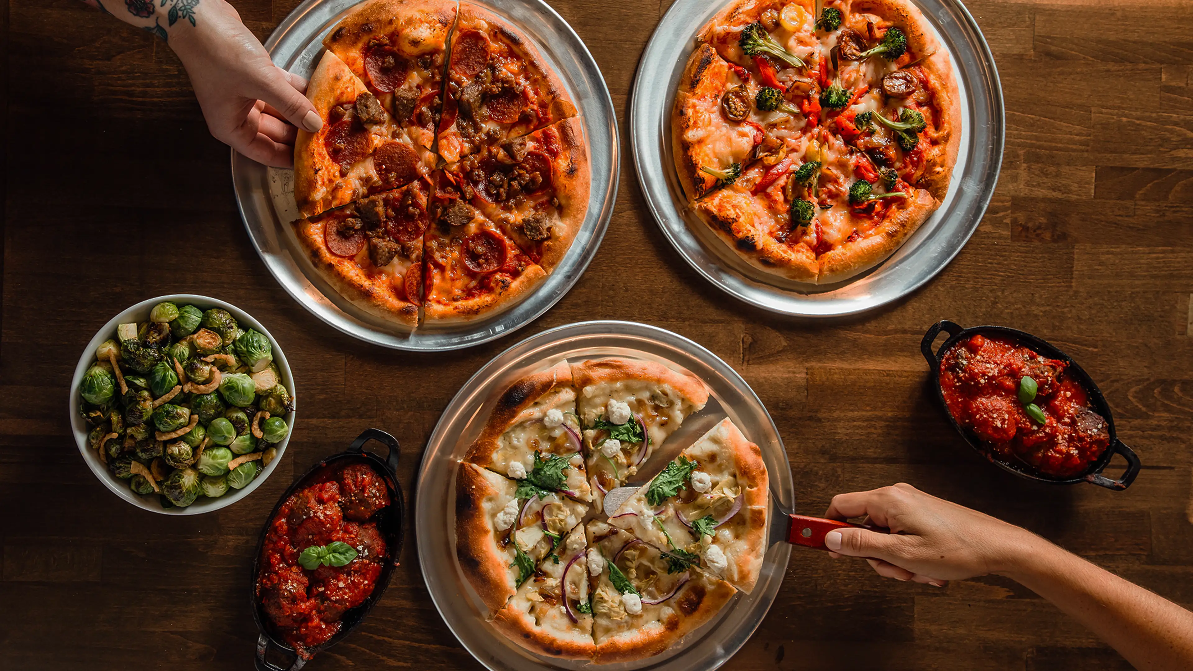 Variety of Wild Pie pizzas on metal trays with side dishes of Brussels sprouts and meatballs in tomato sauce on a wooden table.