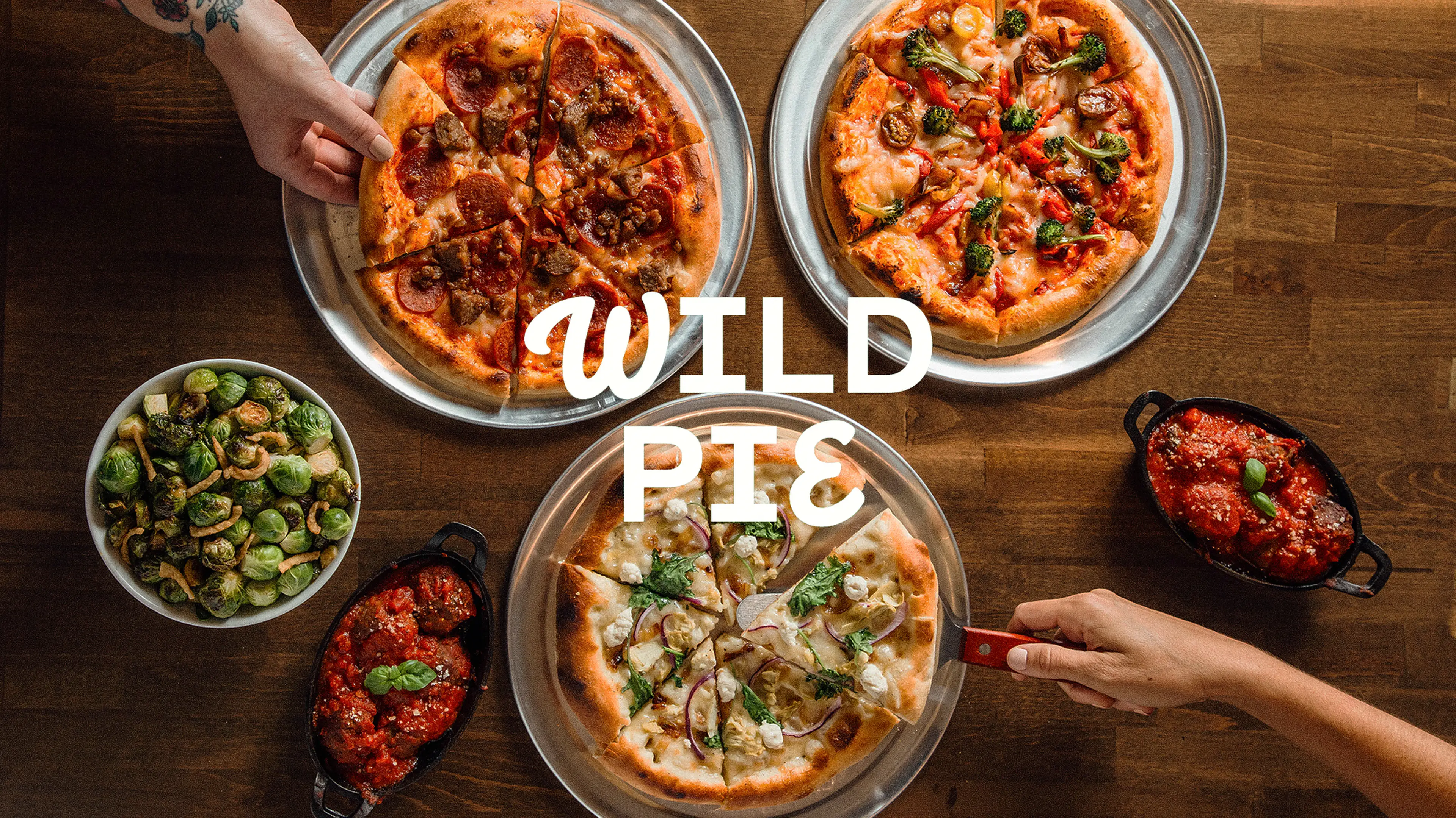 Variety of Wild Pie pizzas on metal trays with side dishes of Brussels sprouts and meatballs in tomato sauce on a wooden table.