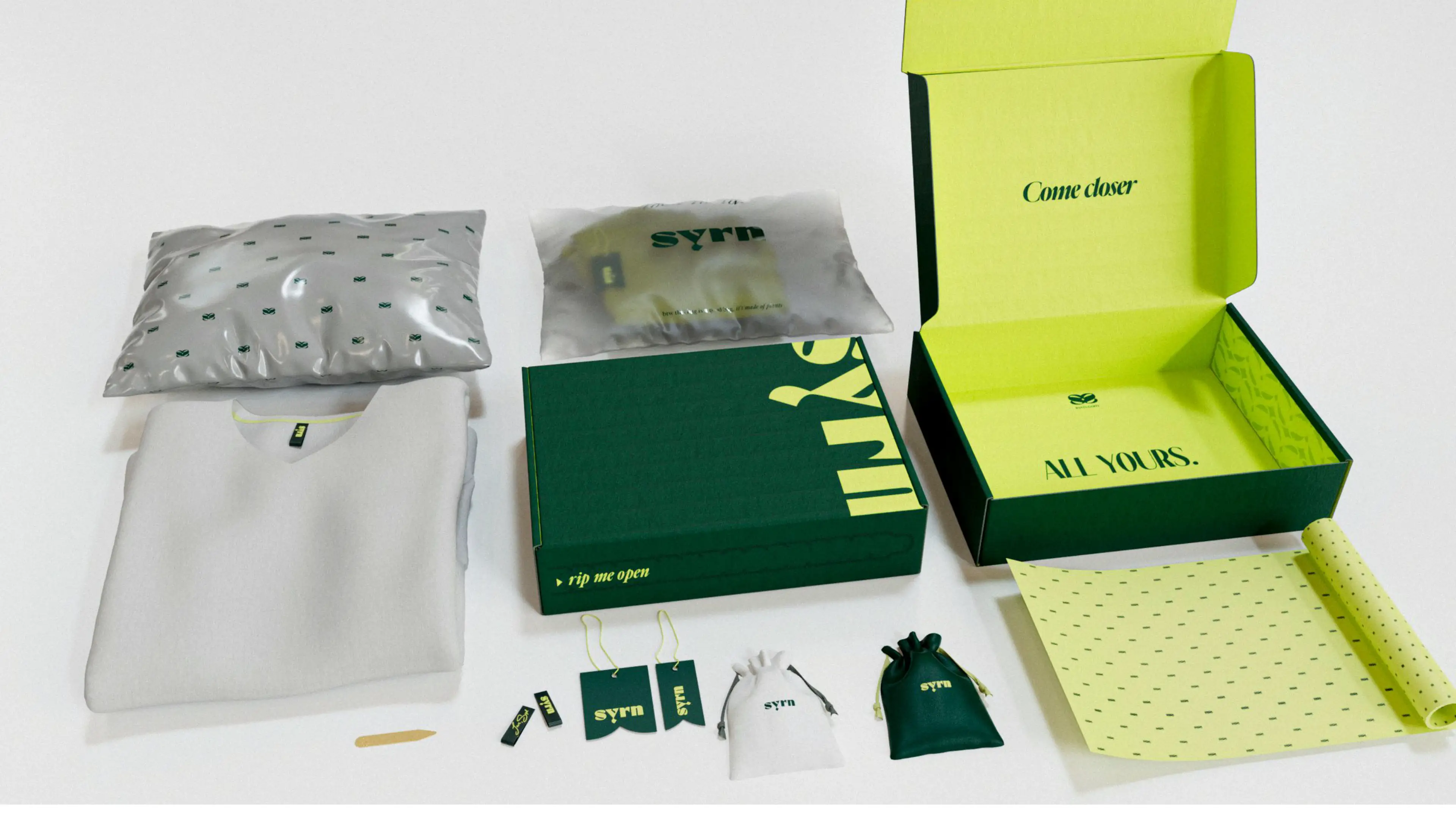 Syrn branded packaging with a folded shirt, sealed bags, boxes, pouches, tags, a wrapping sheet, and a small tool on a white surface.