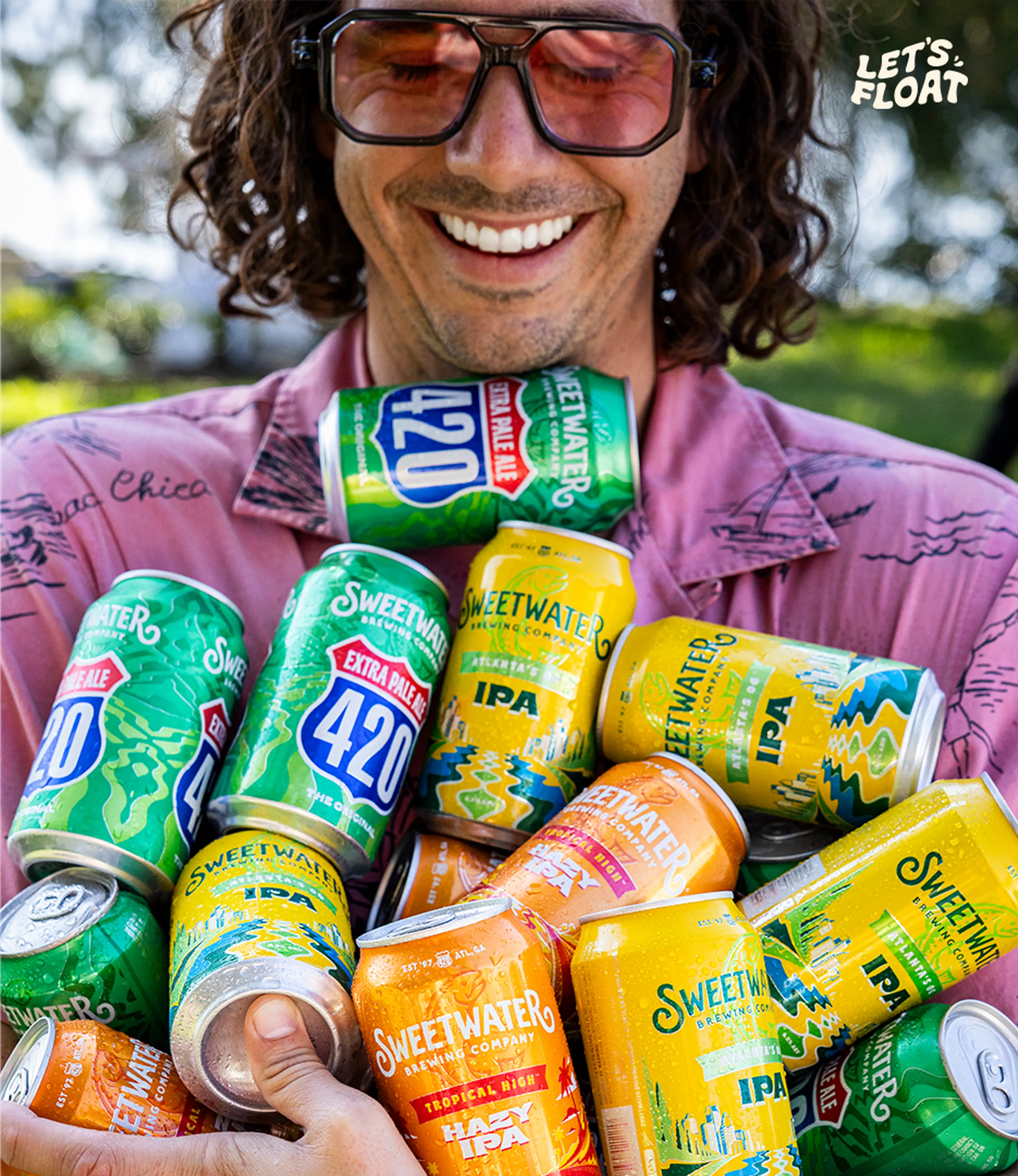Smiling person in sunglasses holding a bundle of colorful SweetWater Brewing Company cans outdoors, with the phrase "Let's Float" in the corner.