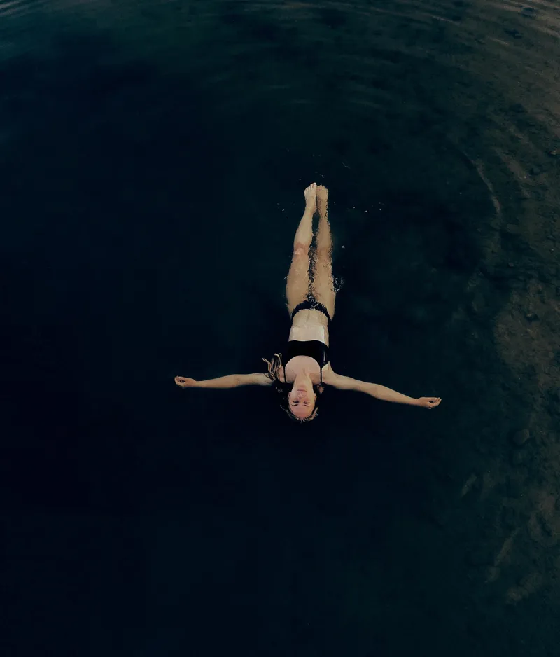Person in a black swimsuit floating on their back in dark, calm water, with arms outstretched and eyes closed, creating a serene atmosphere.