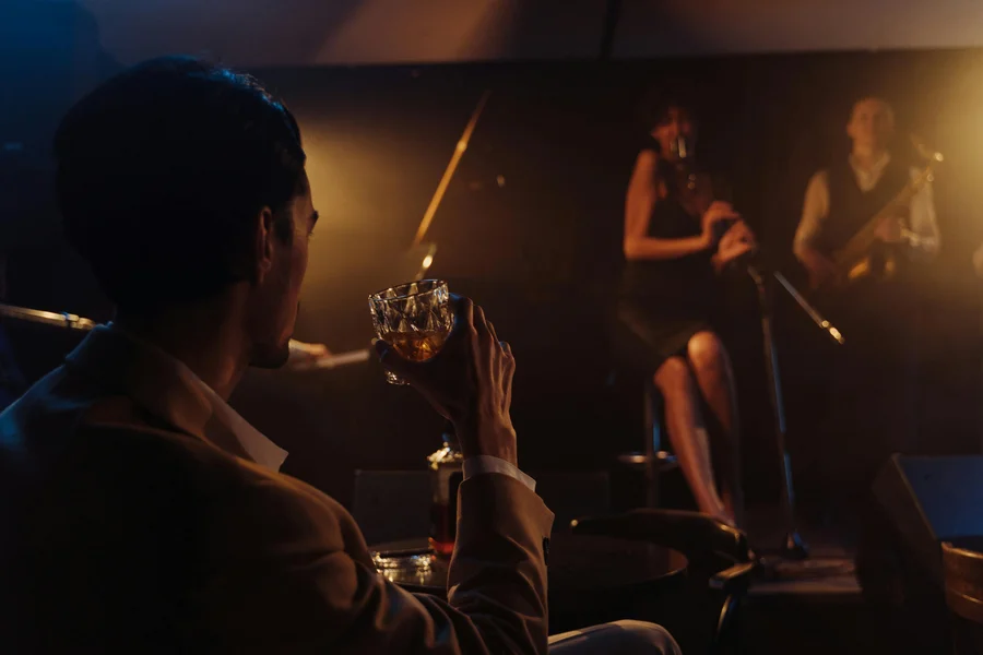 A person holds a drink while watching a woman sing on stage, accompanied by a musician with a guitar. The scene is dimly lit and atmospheric.