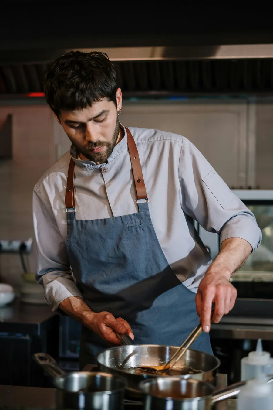 Chef in a kitchen, wearing a blue apron, focused on cooking with a skillet on the stove.