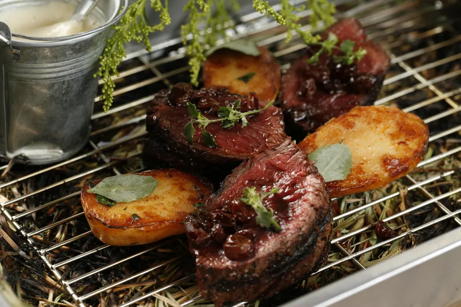 Grilled steak and roasted potatoes garnished with herbs on a metal rack, with a small pot of sauce in the background.