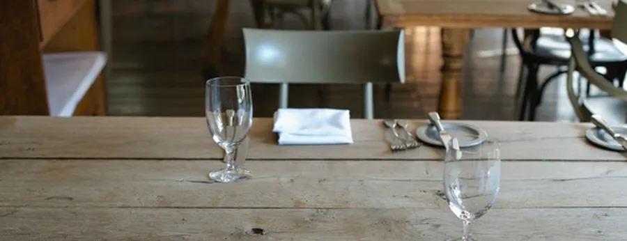 Empty wooden dining table set with two wine glasses, an empty plate, folded napkin, and cutlery in a cozy restaurant setting.