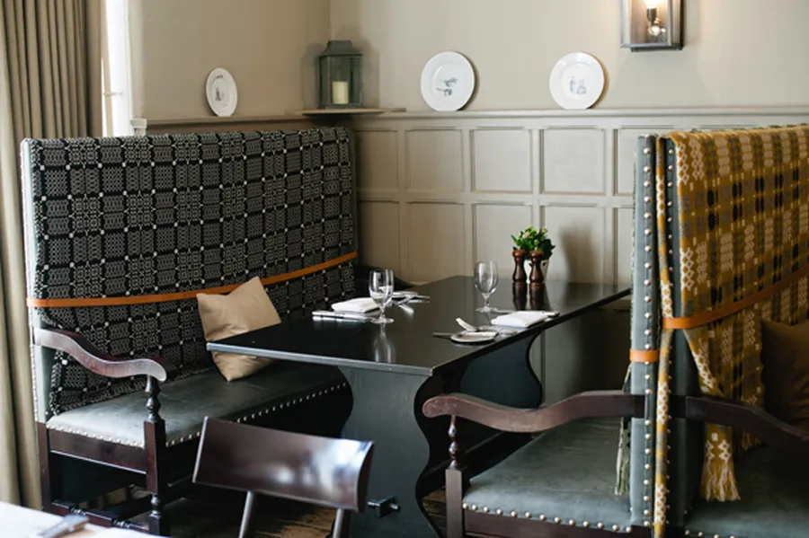 Cozy restaurant booth with patterned high-back seating, set table with wine glasses, cutlery, and a small plant centerpiece against a paneled wall.