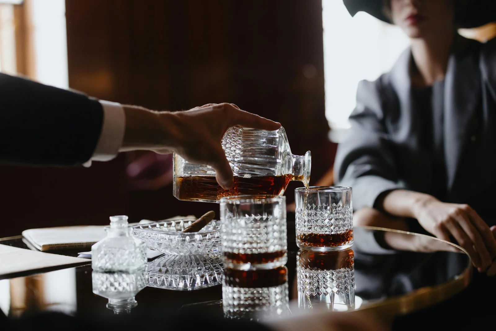 A person pours dark liquor from a decanter into textured glasses on a reflective table, with another person seated nearby in soft focus.