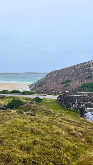 Coastal landscape with a stone bridge, lush green grass, a road, rocky hill, and a sandy beach leading to calm blue water under a cloudy sky.