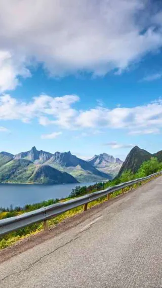 Scenic mountain road with a distant view of peaks and a lake under a partly cloudy blue sky.