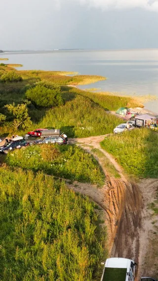 Aerial view of a campsite by a lake with several cars, a tent, and lush greenery, under a cloudy sky.