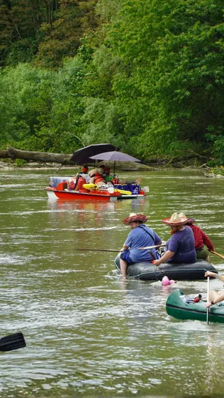 People float down a river on inflatable rafts and a motorized boat with an umbrella, surrounded by dense green foliage.