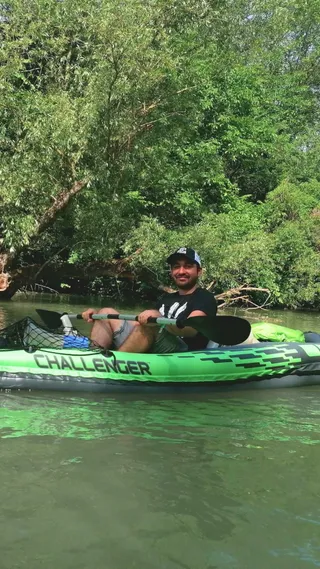 A person in a black shirt and cap paddles a green inflatable kayak on a calm river surrounded by lush green trees.