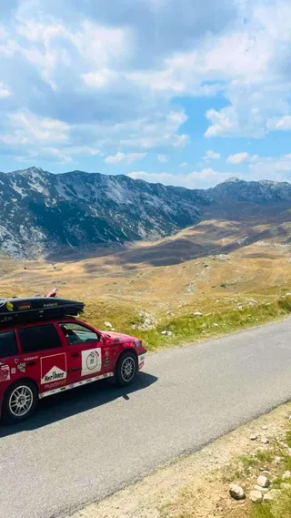 Red car on a mountain road with distant peaks under a blue sky and scattered clouds.