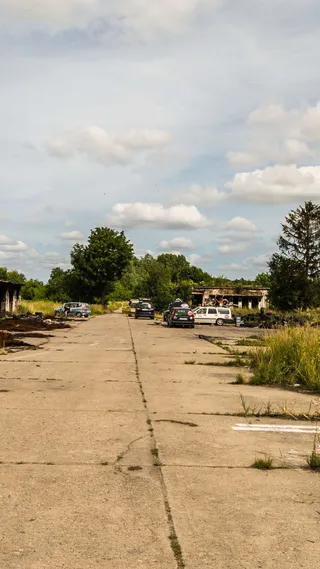 Abandoned concrete road with scattered cars and overgrown vegetation, under a partly cloudy sky.