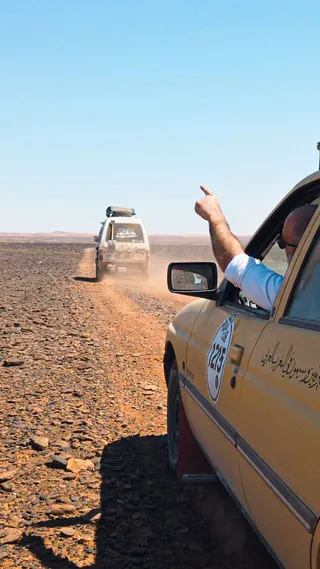 Two vehicles driving on a rocky desert road, with a man in the nearest car pointing ahead, under a clear blue sky.