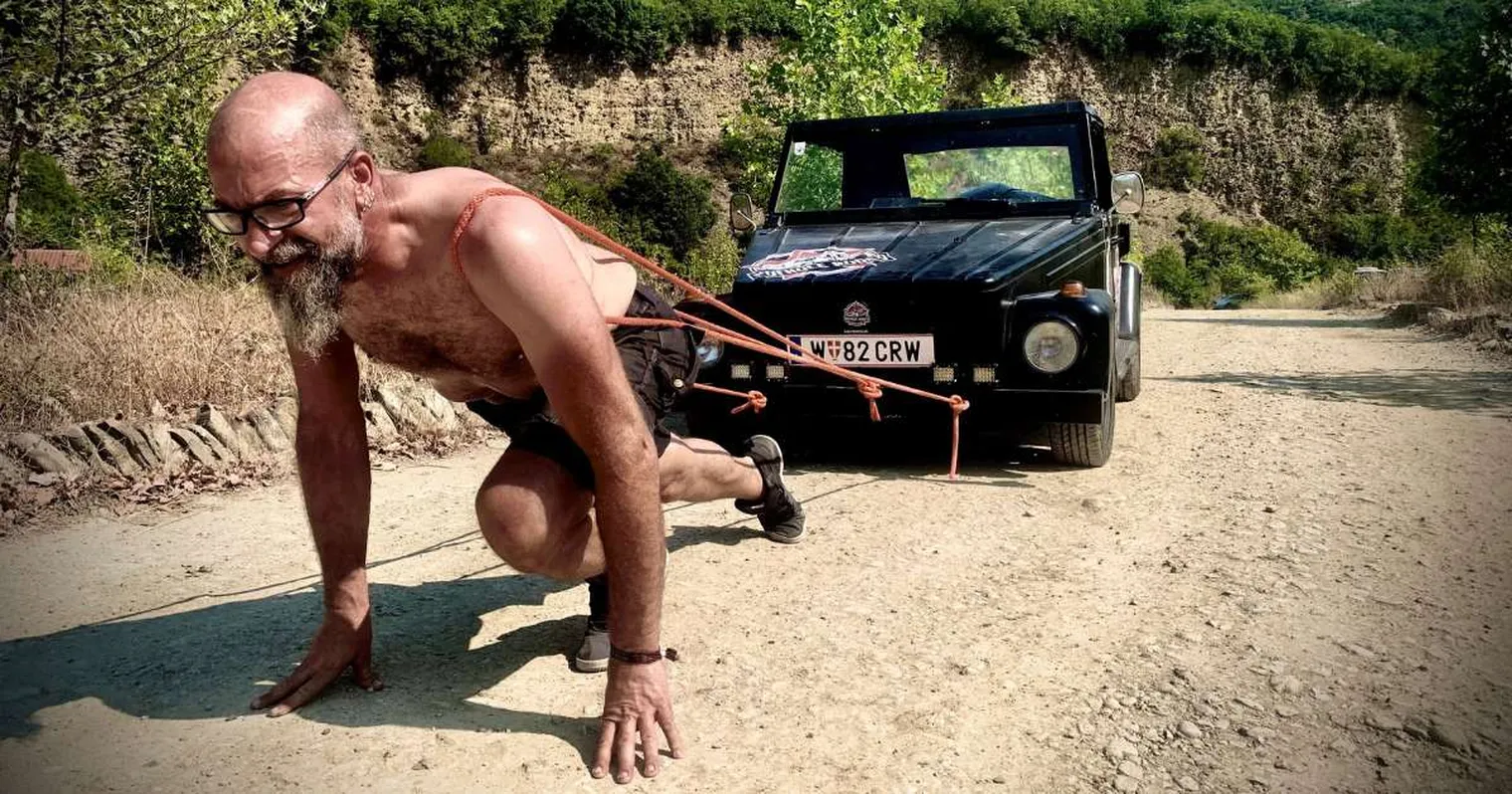A shirtless man, wearing glasses, is pulling a black car using ropes on a dirt road surrounded by trees and rocky terrain.