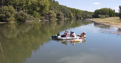 People on a raft and paddleboard on a calm river with lush green trees and a clear sky reflecting in the water.