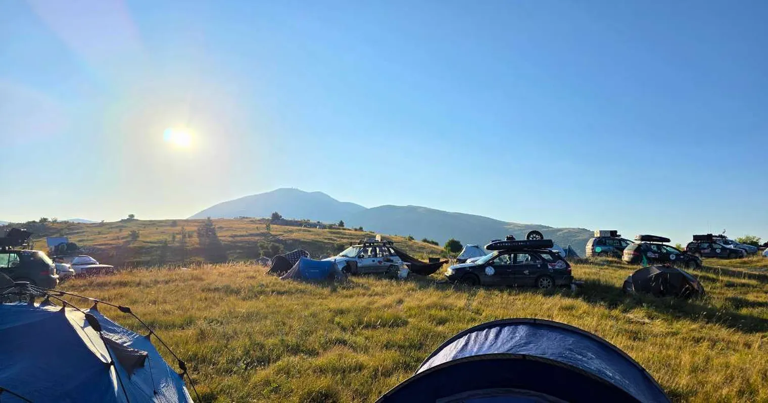 Cars and tents scattered on a grassy hillside with a mountain in the background, under a clear blue sky and bright sun.