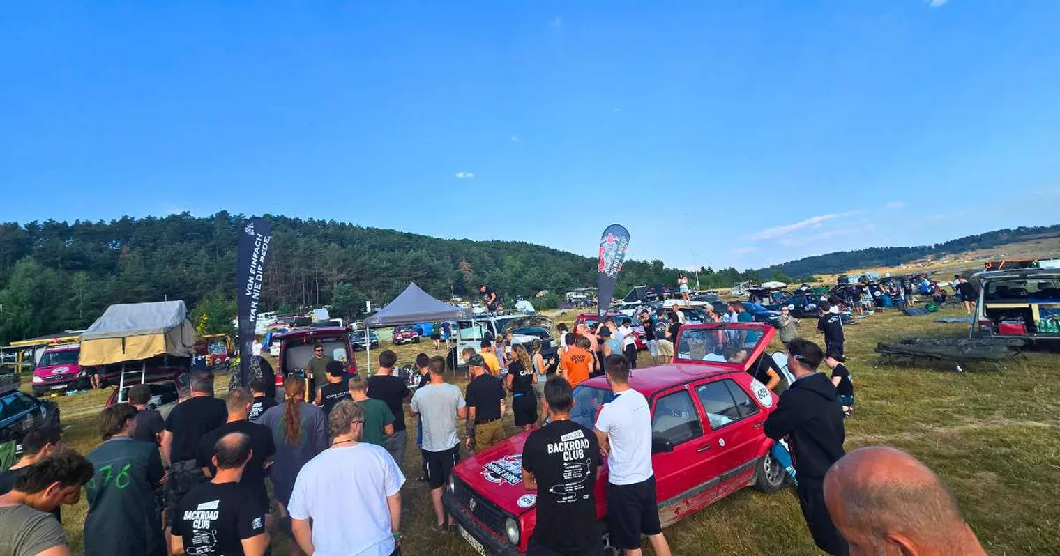 A crowd gathers around a red car at an outdoor event with tents and cars on a grassy field under a clear blue sky.