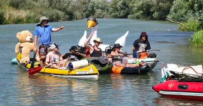 Group of people in hats on inflatable rafts with a giant teddy bear and swan float, enjoying a sunny day on a river.