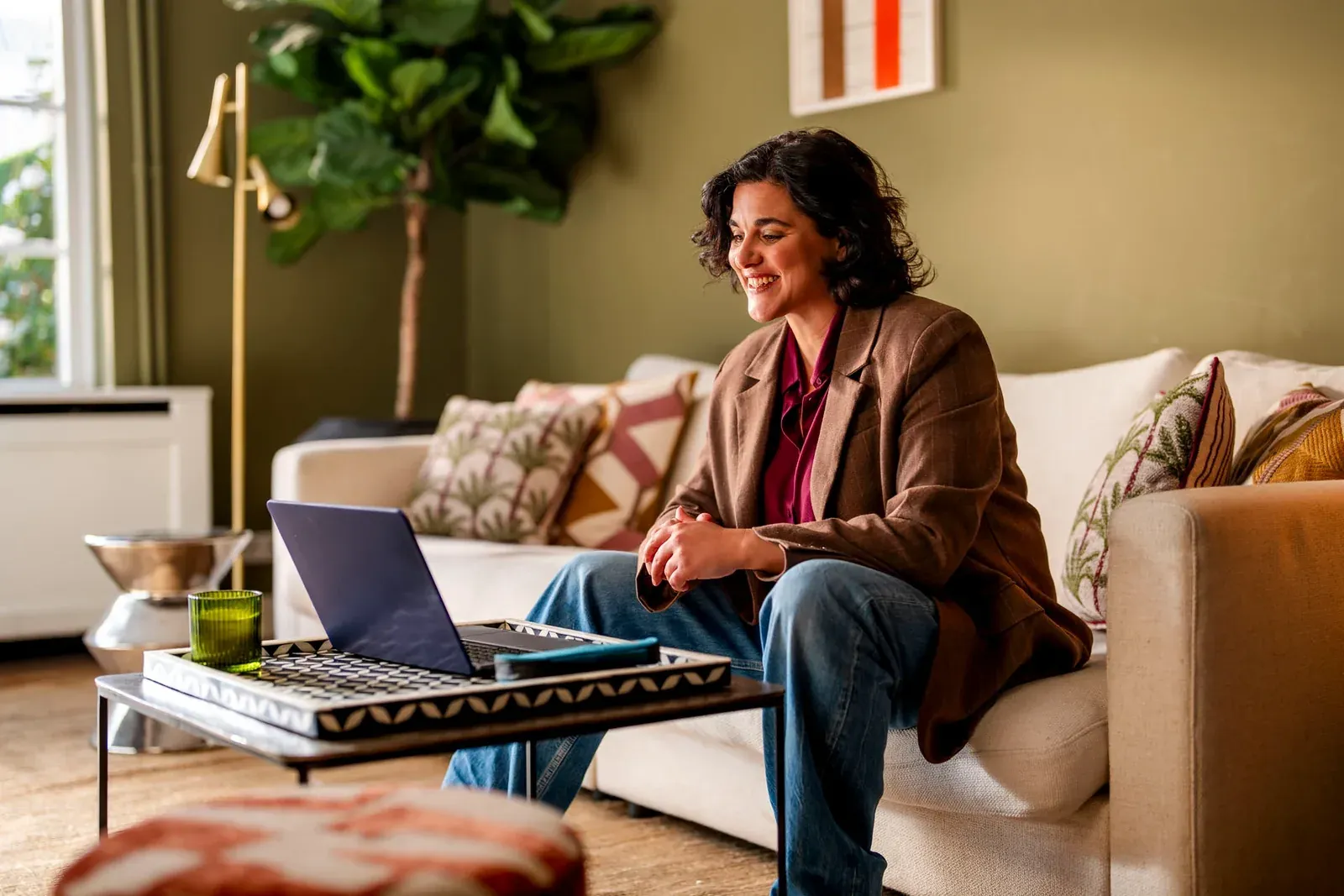 Vrouw in een bruin jasje zit op de bank met een laptop voor zich