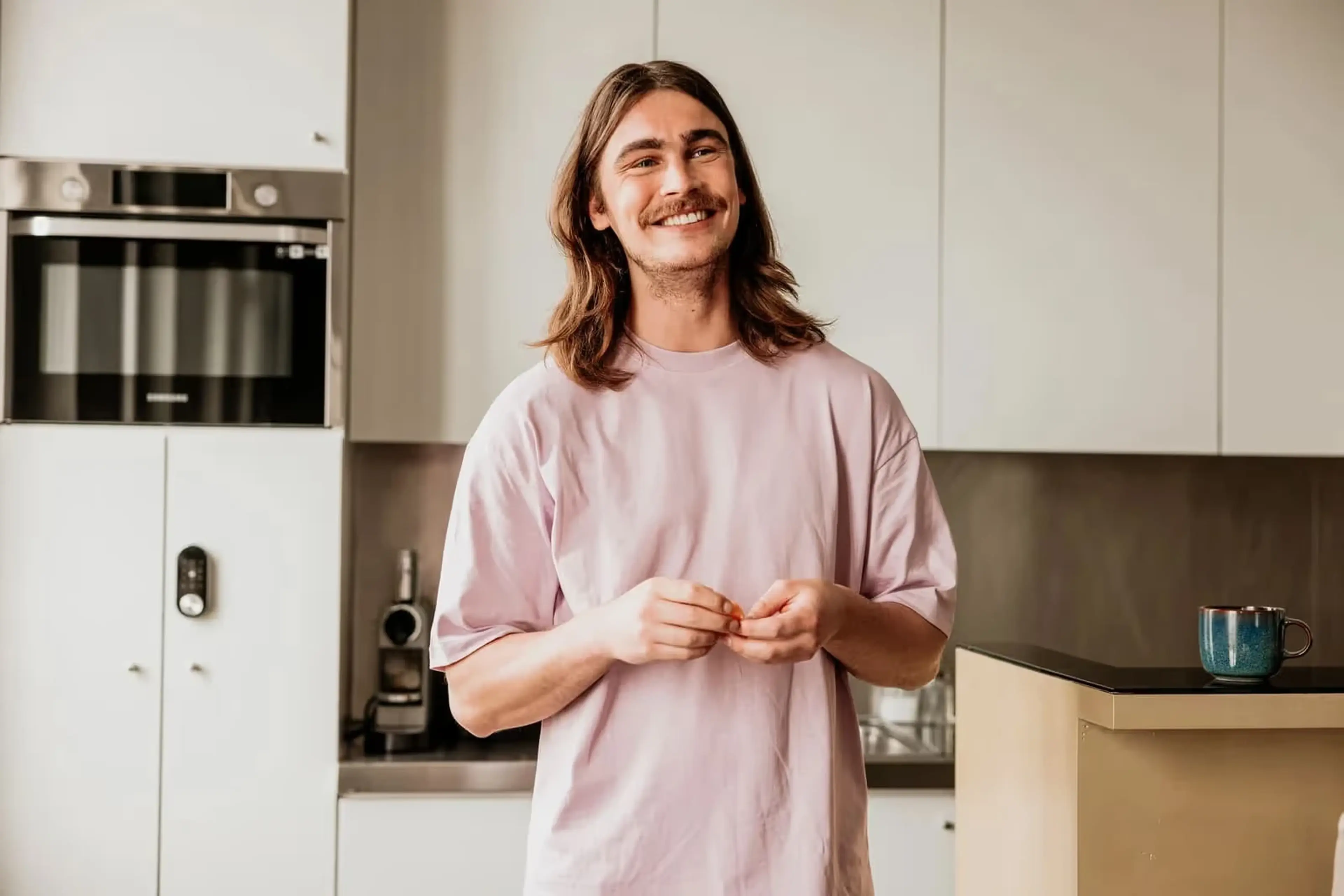 Lachende man met lang haar staat ontspannen in zijn lichte keuken