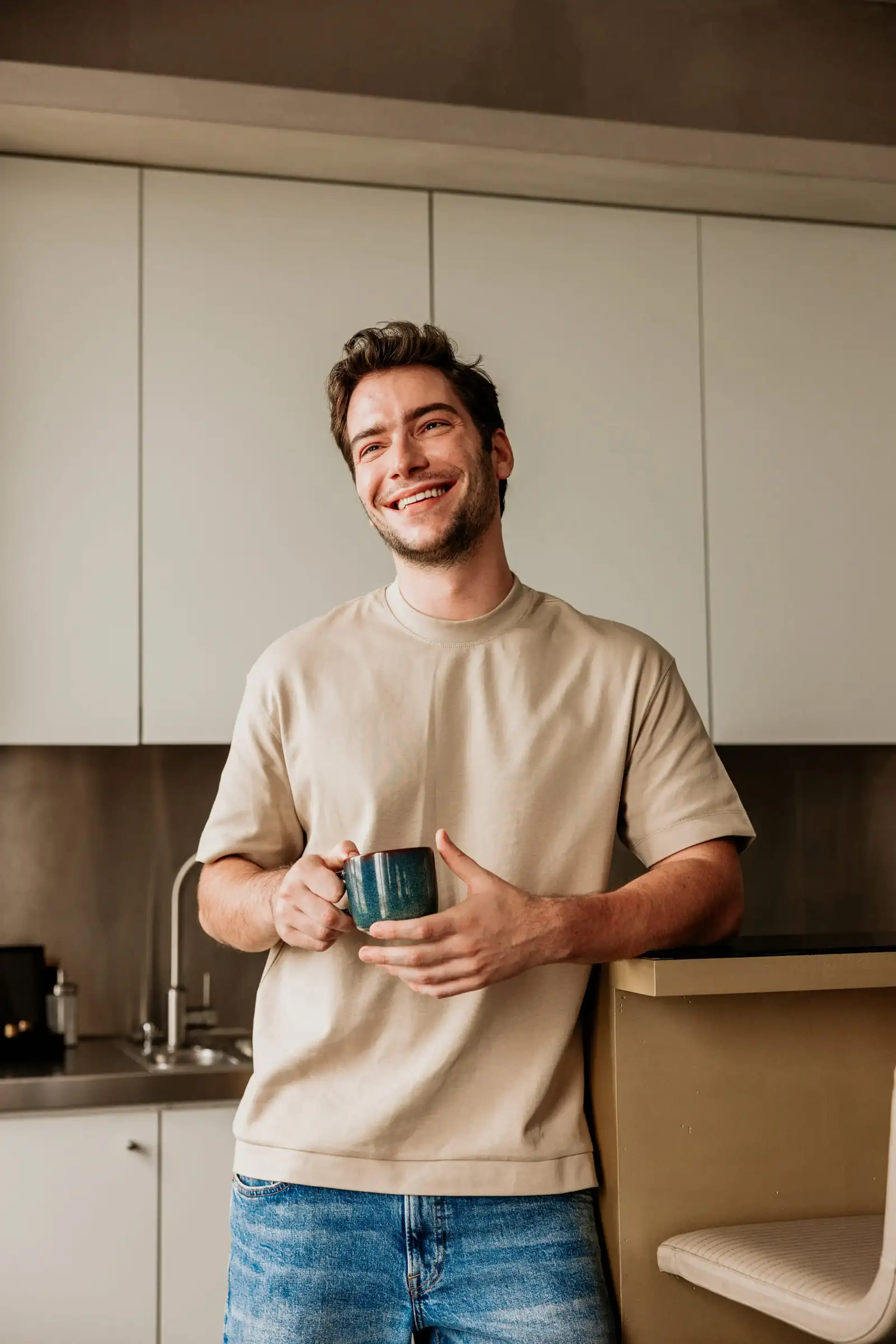 Lachende man in een beige T-shirt staat ontspannen in een lichte keuken met een blauwgroene mok in zijn handen.