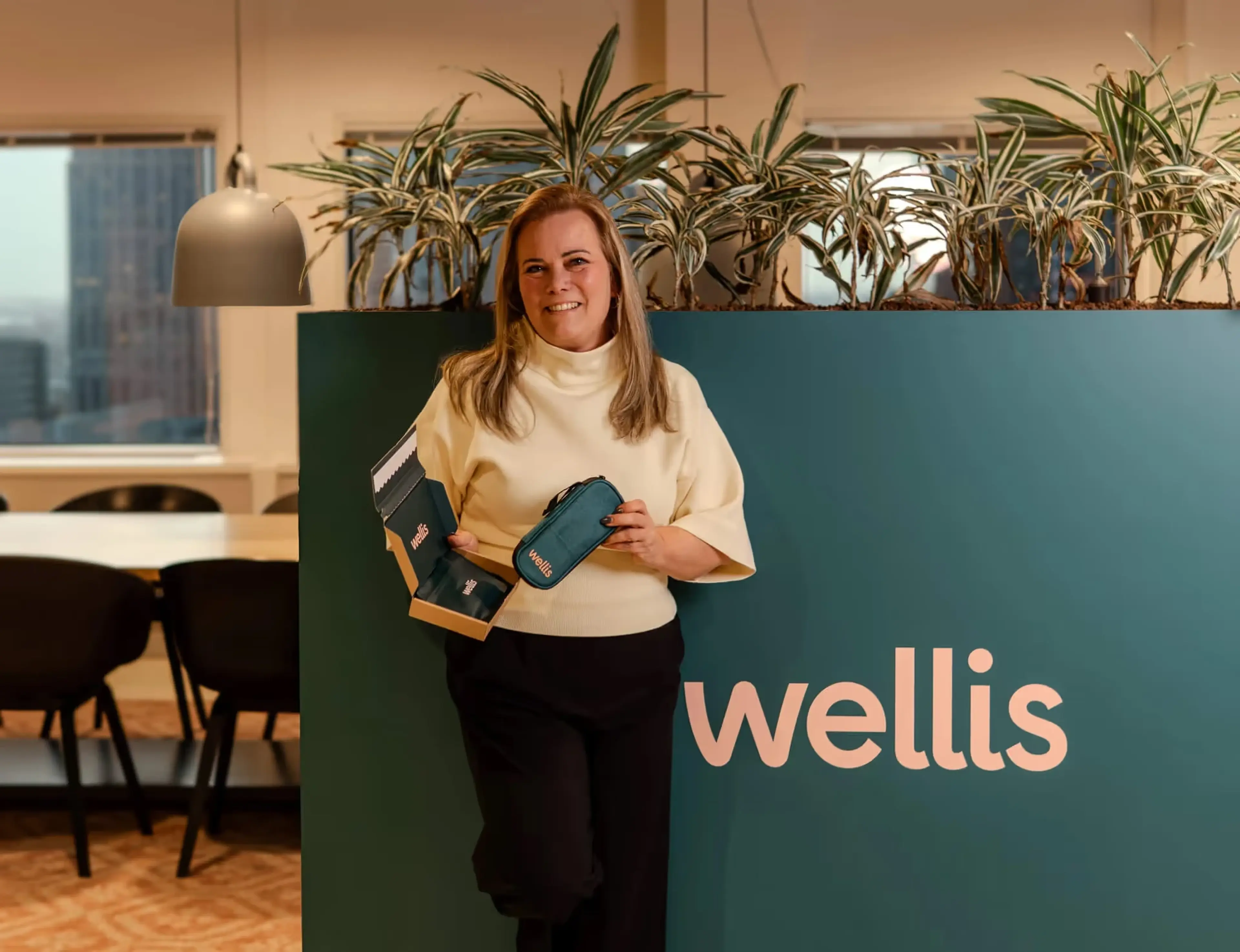 Eveline Engels staat lachend voor een groene wand met het Wellis-logo, met een geopende Wellis-doos en een blauwgroen Wellis-etui in haar handen.