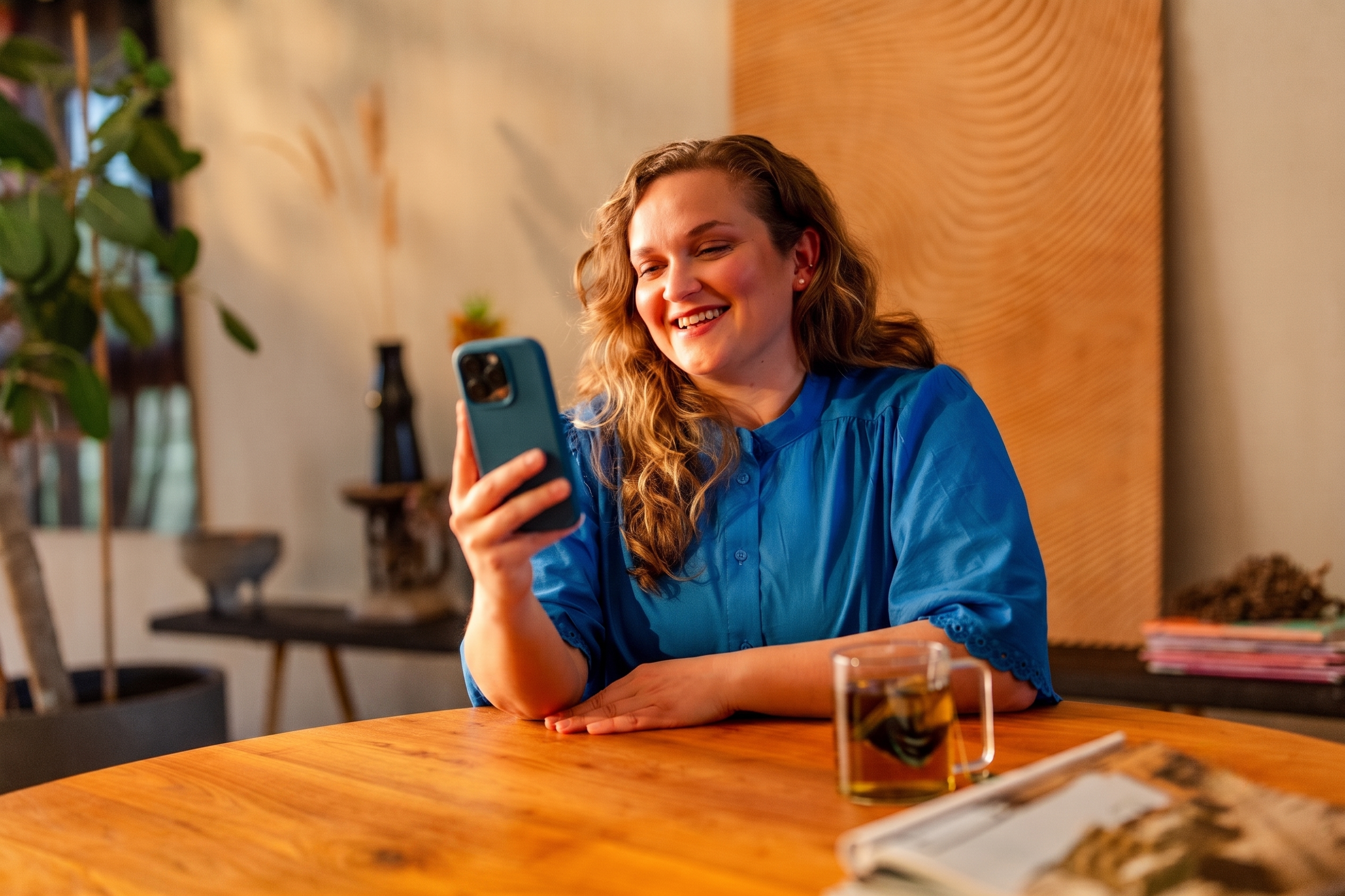 Lachende vrouw zit aan een houten tafel met haar telefoon in de hand en een glas kruidenthee binnen handbereik.