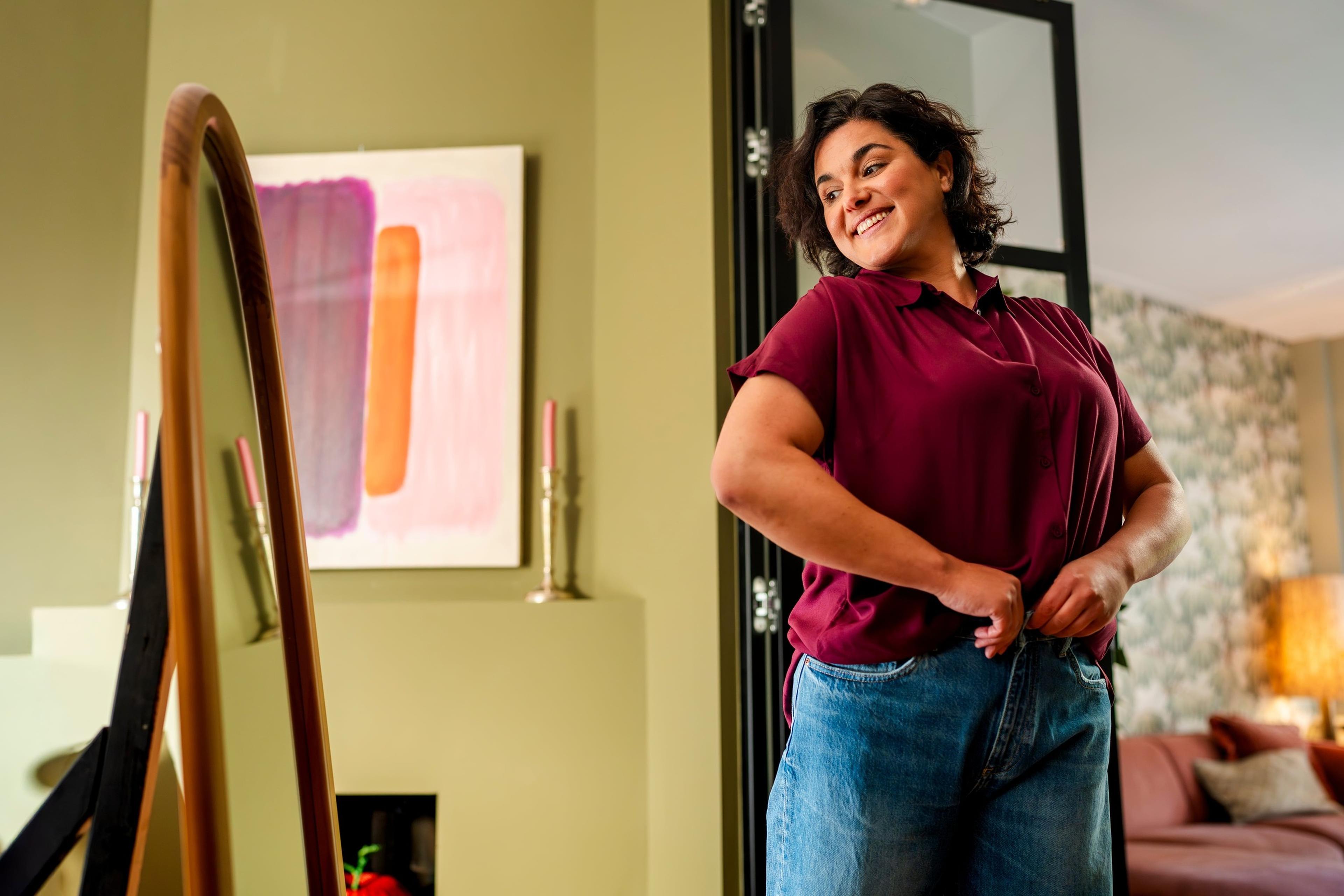 Lachende vrouw stopt haar bordeauxrode blouse in haar spijkerbroek voor een grote staande spiegel in de woonkamer.