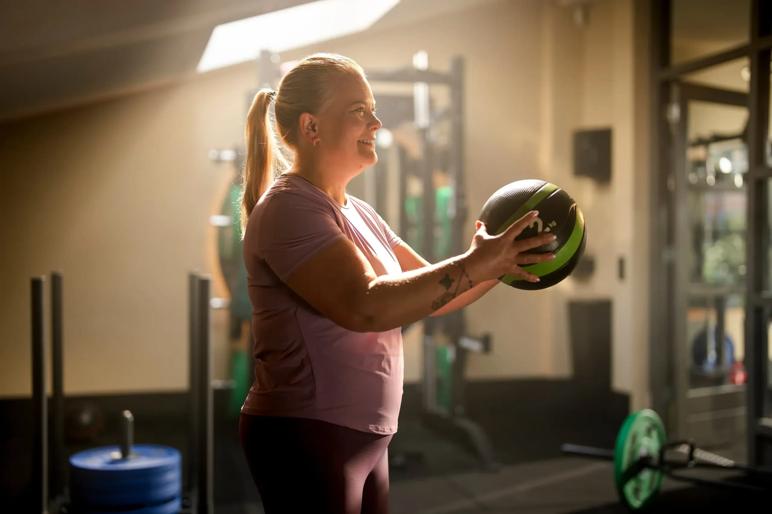 Vrouw in sportkleding houdt een medicijnbal voor haar borst in een sportschool met halterrek en gewichtsschijven.