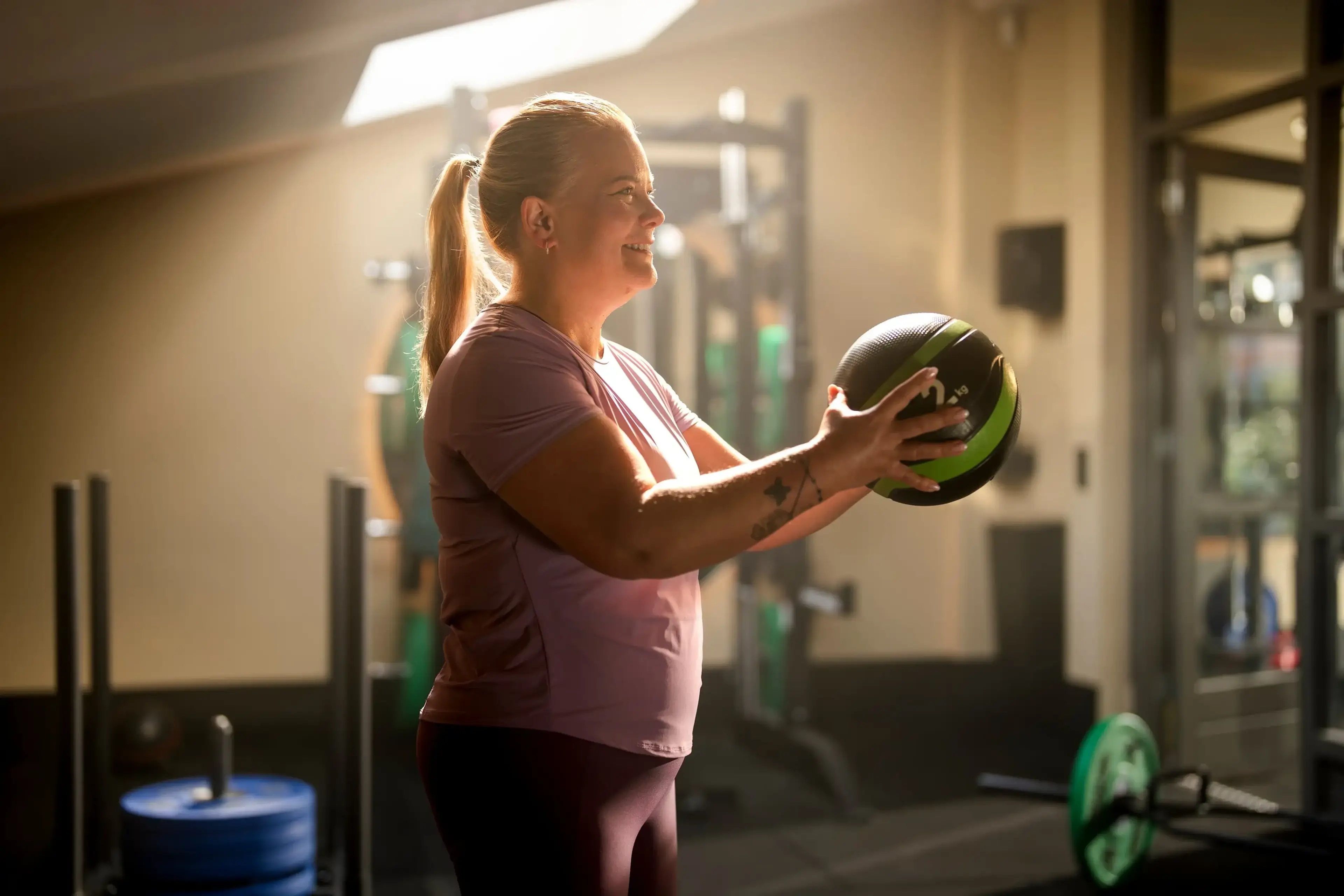 Vrouw in sportkleding houdt een medicijnbal voor haar borst in een sportschool met halterrek en gewichtsschijven.