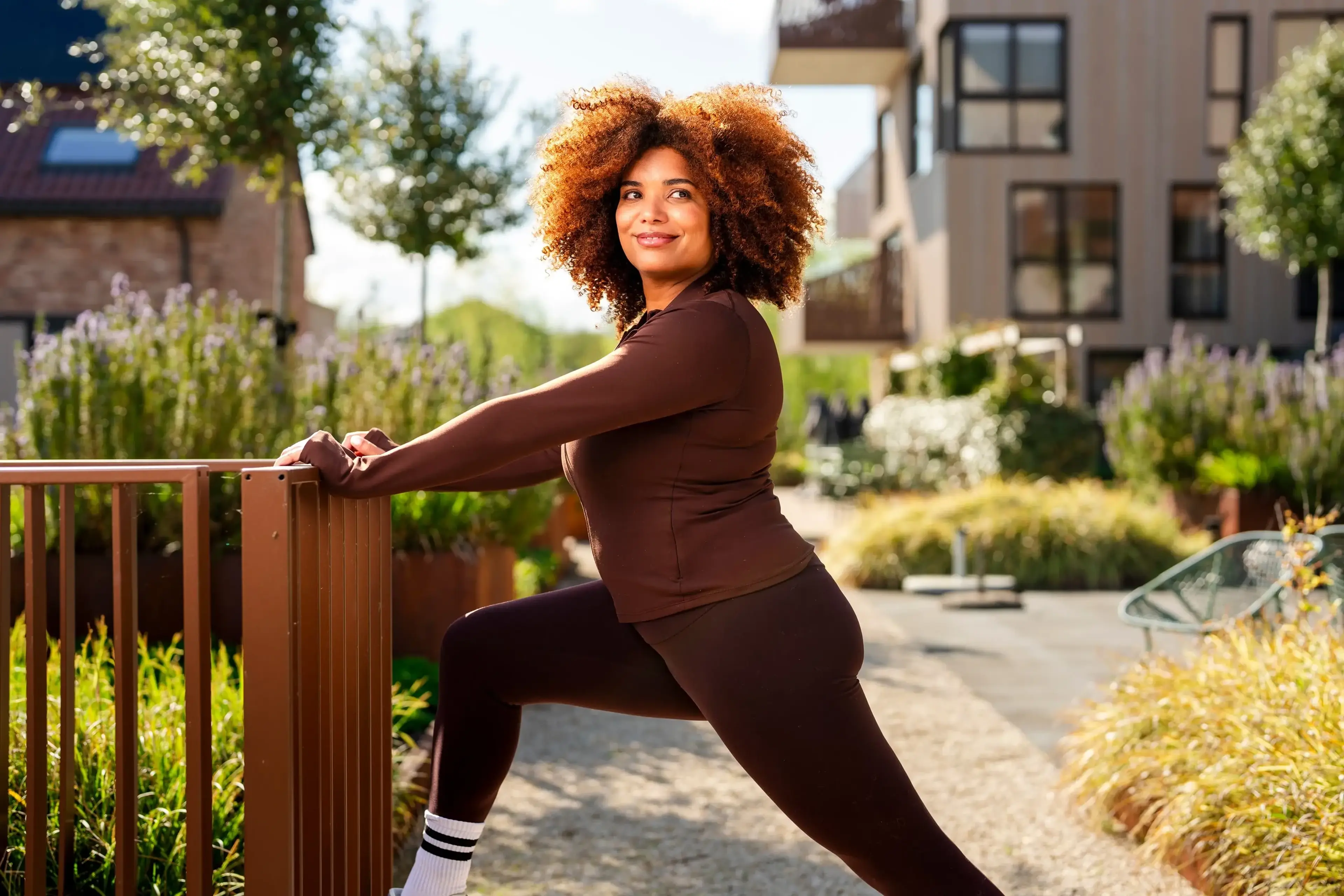 Lachende vrouw in bruine sportkleding rekt haar been uit tegen een houten hek in een zonnige binnentuin.