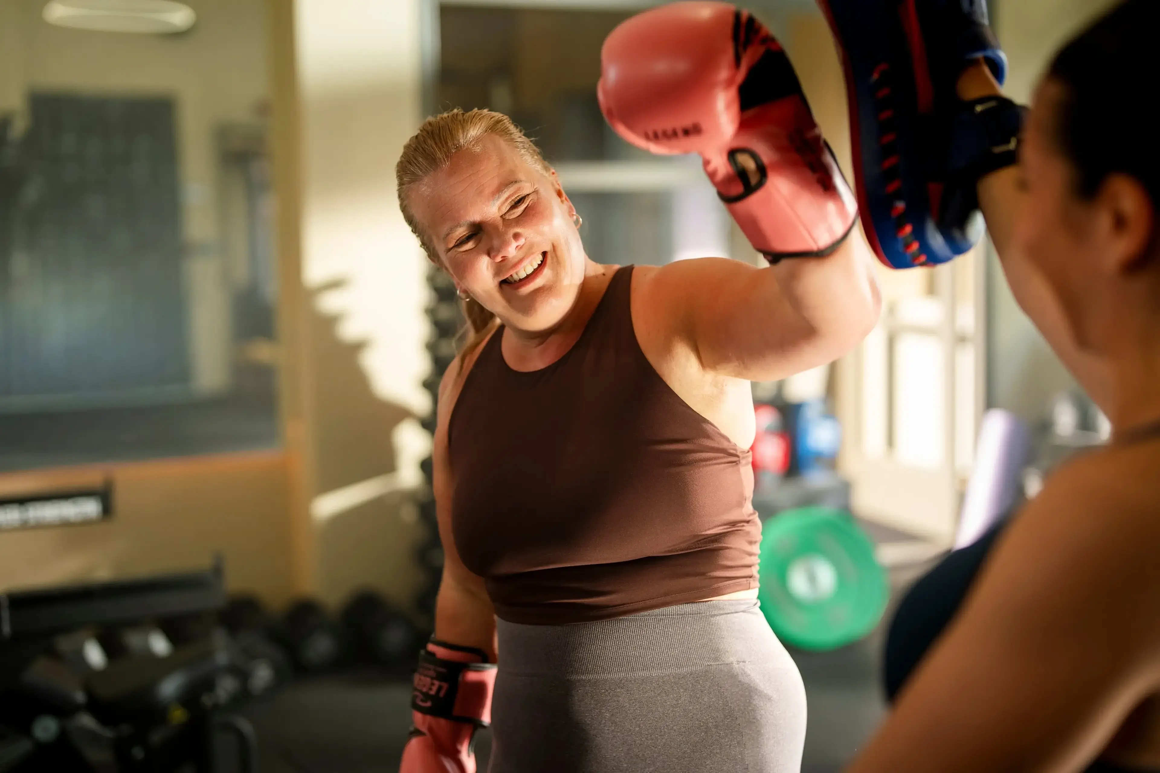 Lachende vrouw met roze bokshandschoenen slaat een stoot op de handpads van haar trainer in de sportschool.