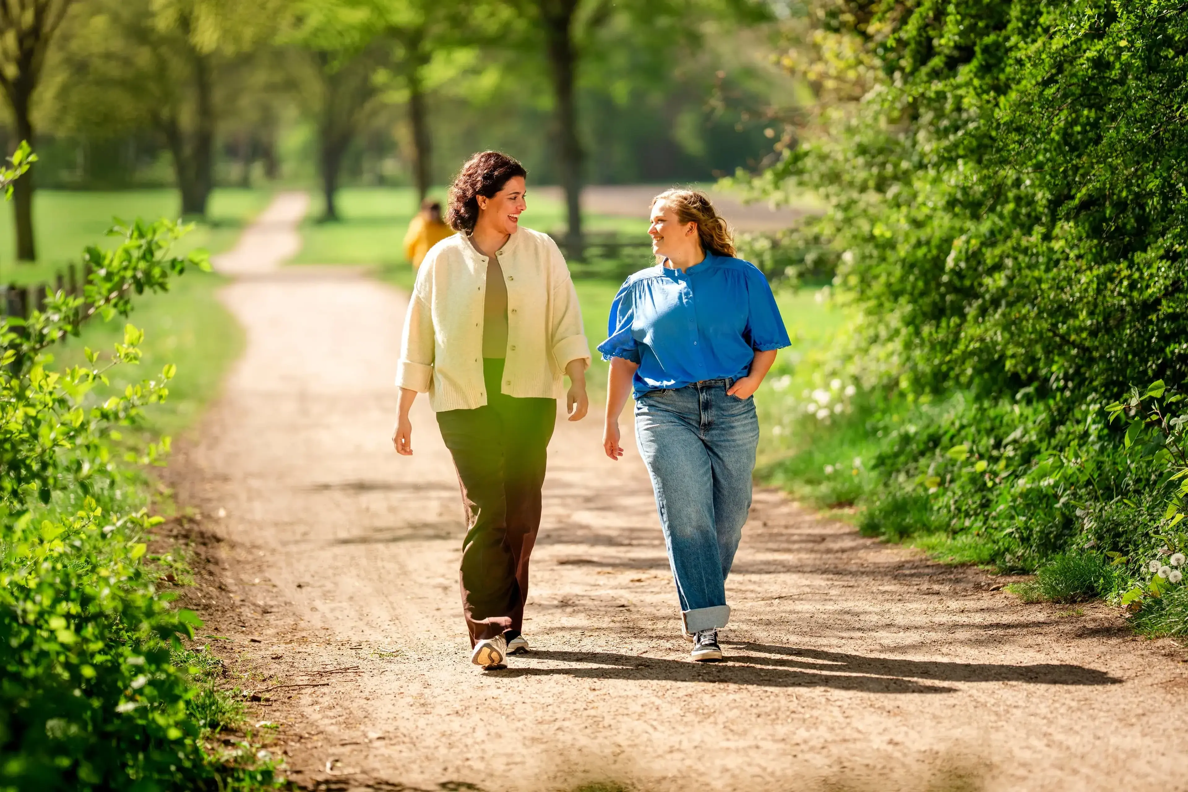 Twee vrouwen wandelen pratend naast elkaar over een zandpad door een groen voorjaarspark.