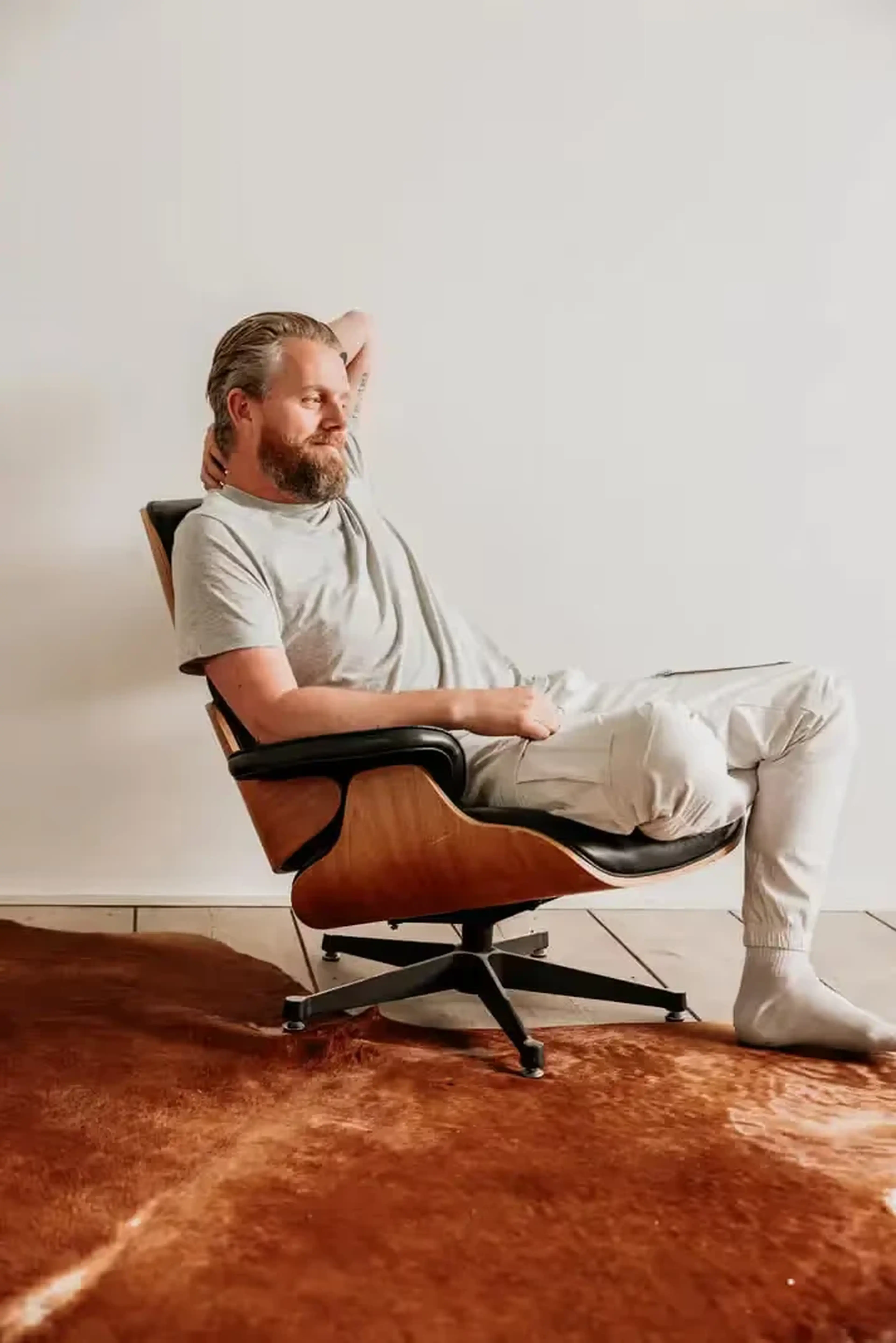Ontspannen man met baard leunt met zijn hand achter zijn hoofd achterover in een leren loungestoel tegen een witte muur