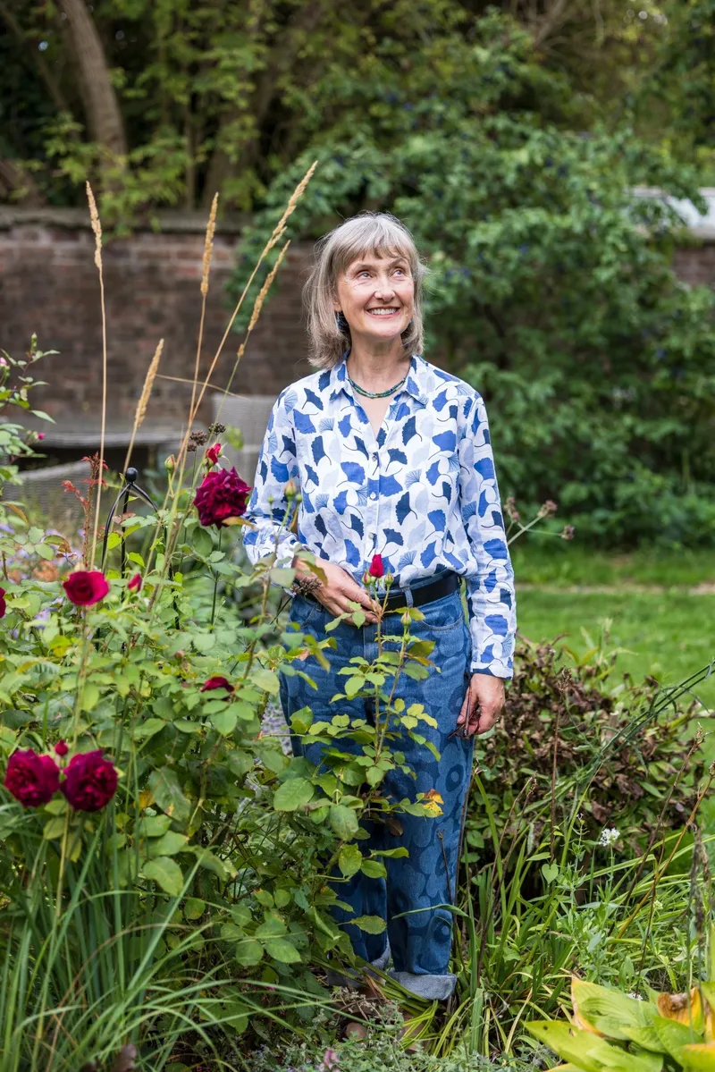 Sally Tierney smiling and looking up, standing among dark red roses and tall grasses in a walled garden