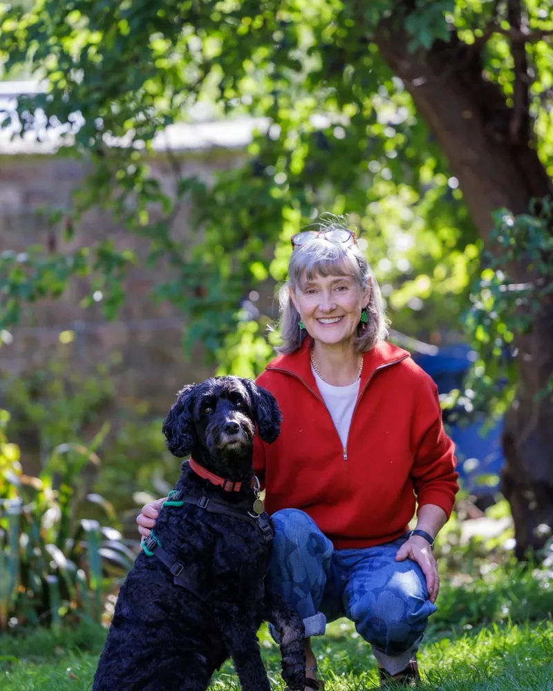 Sally Tierney crouching on a lawn beside Kibby, her black cockapoo, under a tree in a walled garden