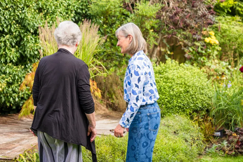 Sally Tierney walking round a garden with its owner, who is seen from behind, looking at the planting beside a timber deck